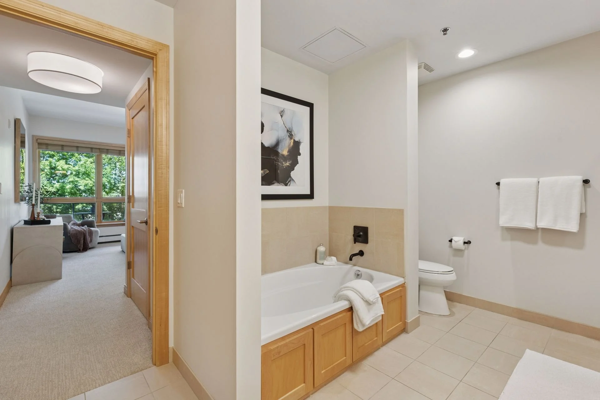 A bathroom with a bathtub, a toilet, and towels on a rack, with a partial view of a living room with large window and furniture in the background.