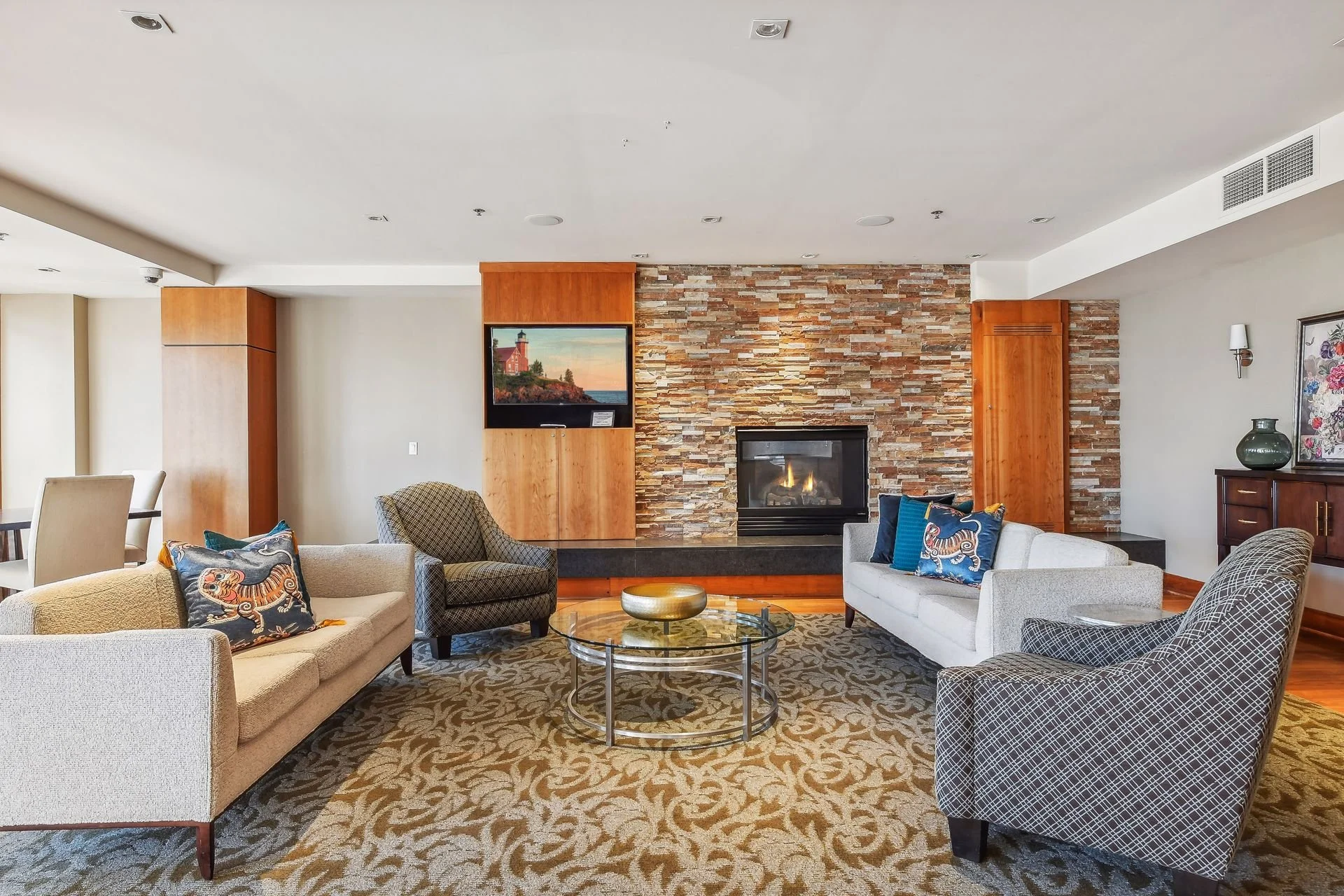 Community room featuring a stone fireplace, two white sofas with colorful throw pillows, two patterned armchairs, a glass coffee table with a decorative bowl, a wall-mounted TV, and a patterned area rug.