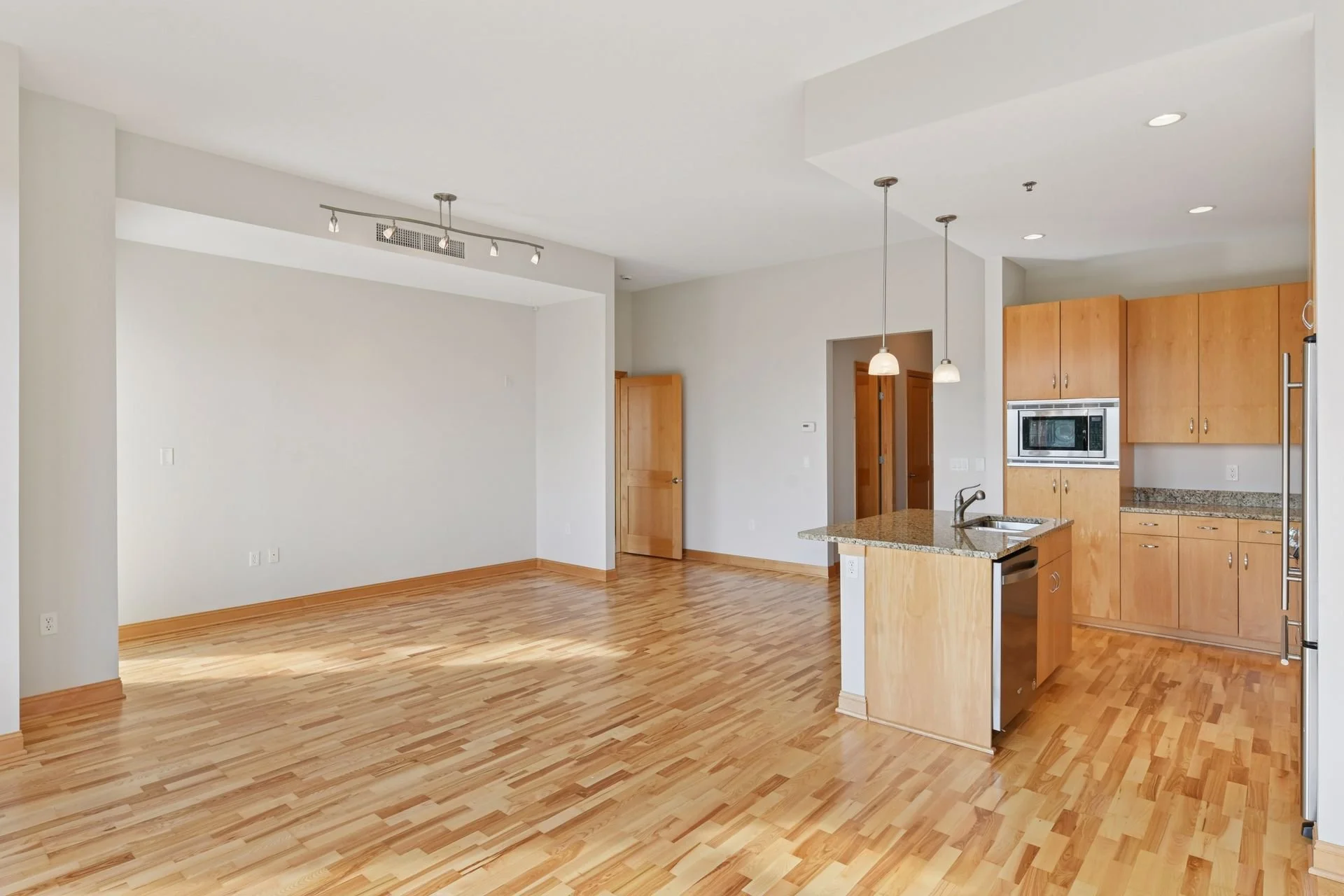 Empty open-concept living area and kitchen with hardwood floors, white walls, wooden cabinets, granite countertops, and ceiling lighting fixtures.