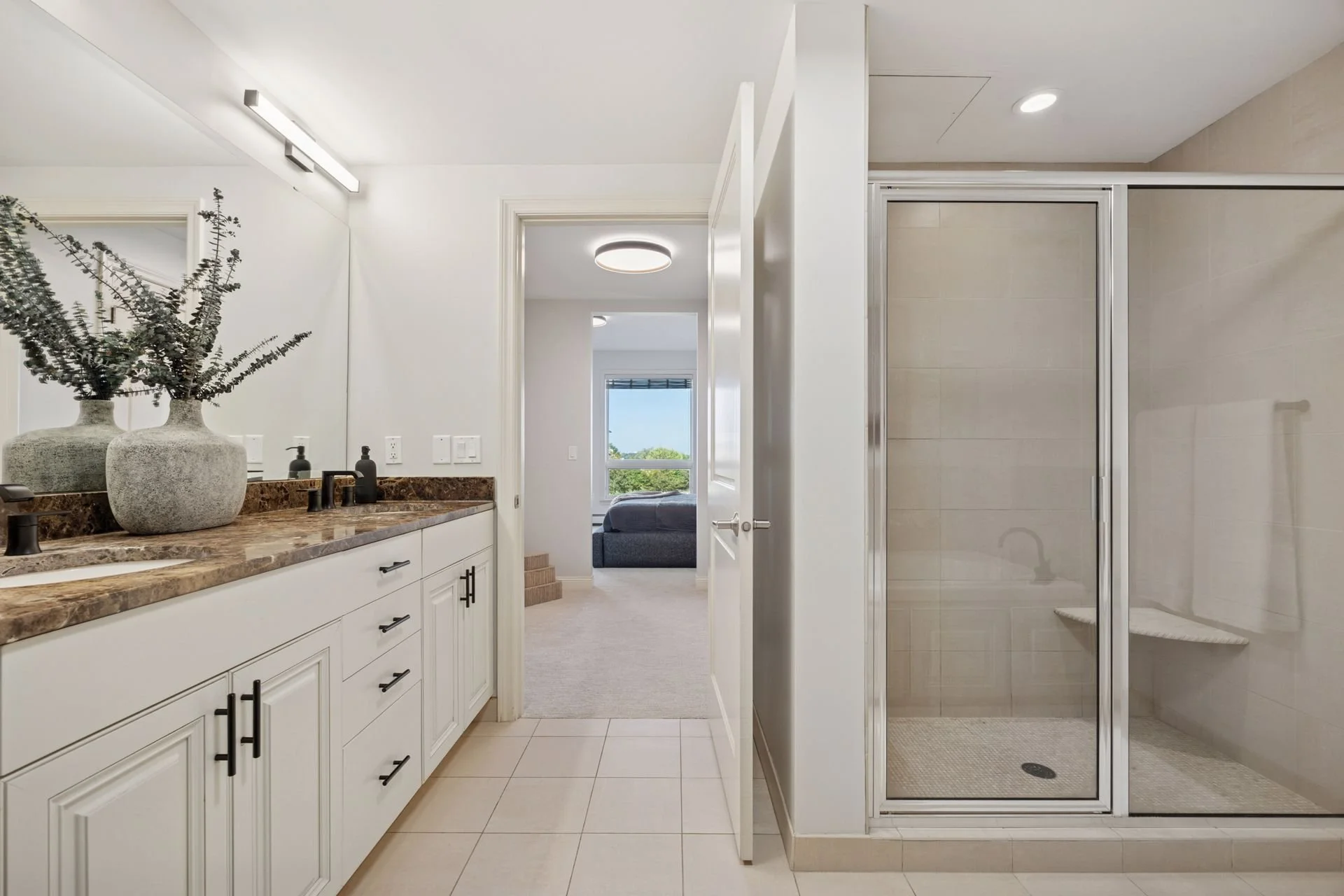 Bathroom with white cabinets, brown marble countertop, and black fixtures; large mirror; glass shower with beige tiles; doorway leading to a bedroom with a window view of greenery.