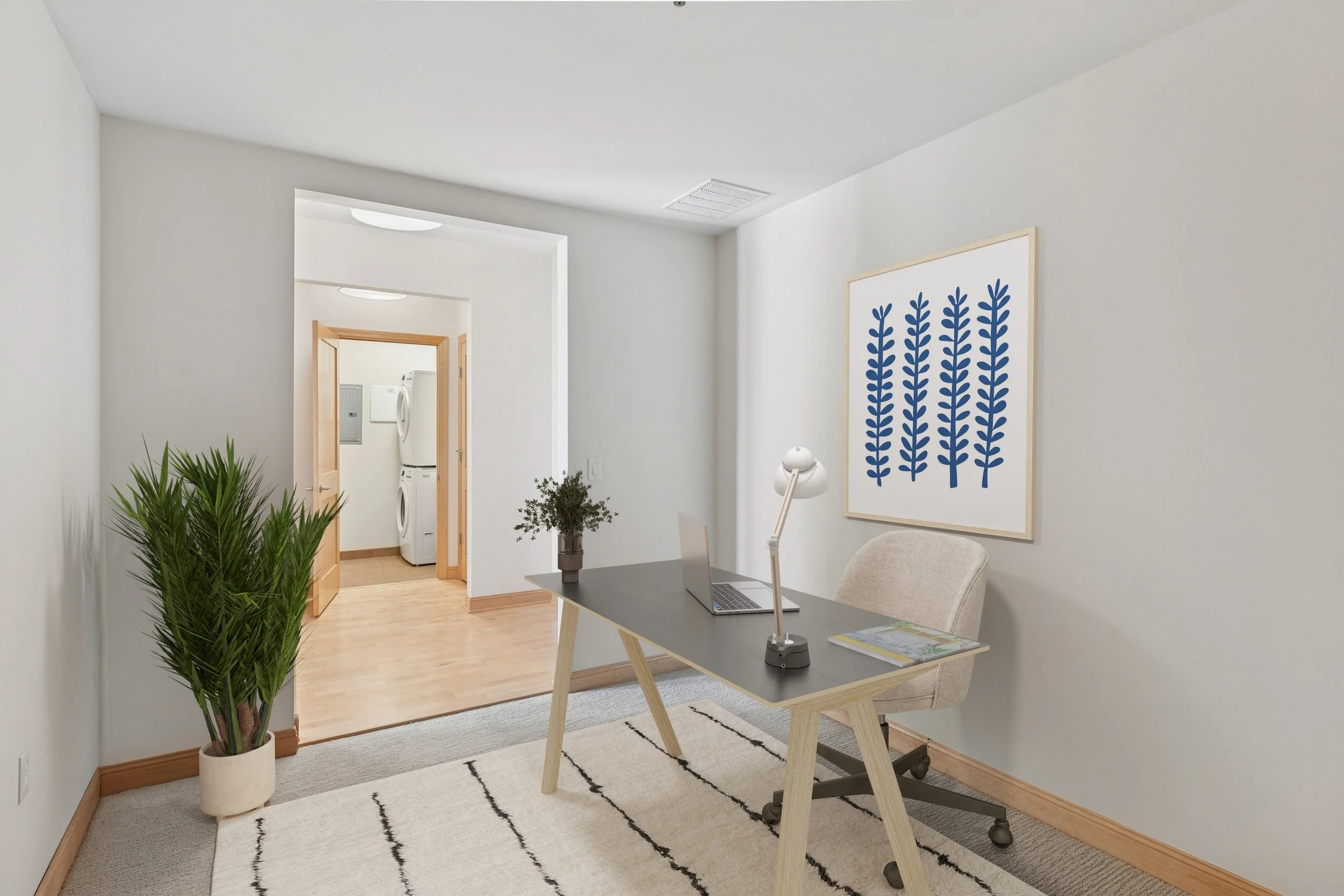 Home office with desk, laptop, lamp, framed blue leaf artwork, beige chair, potted plant, and laundry area in the background