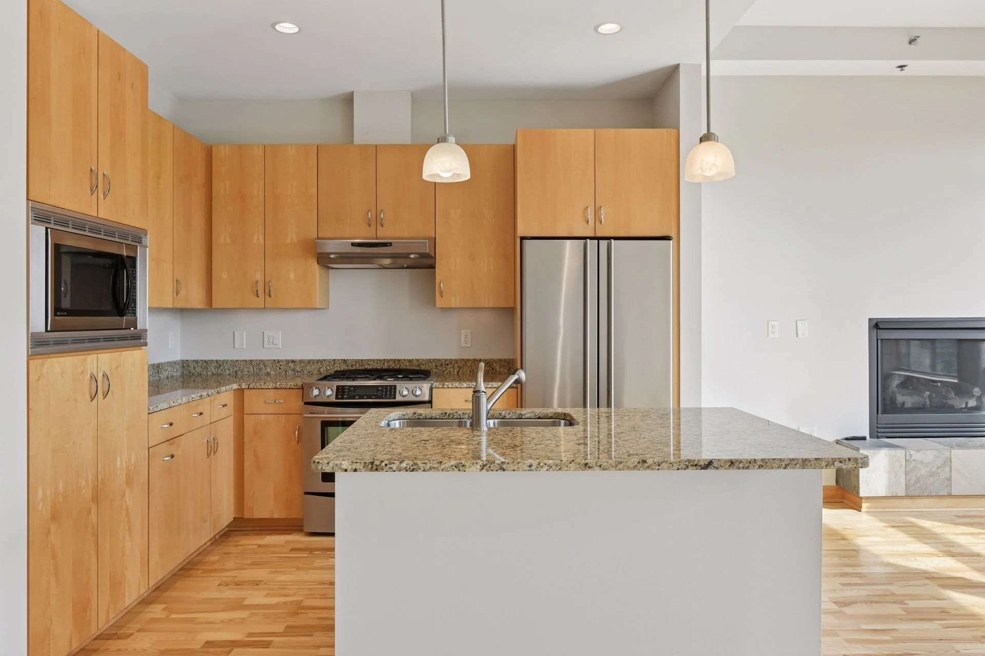 Modern kitchen with wooden cabinets, granite countertops, stainless steel appliances, and a small island with a sink, in an open living space.