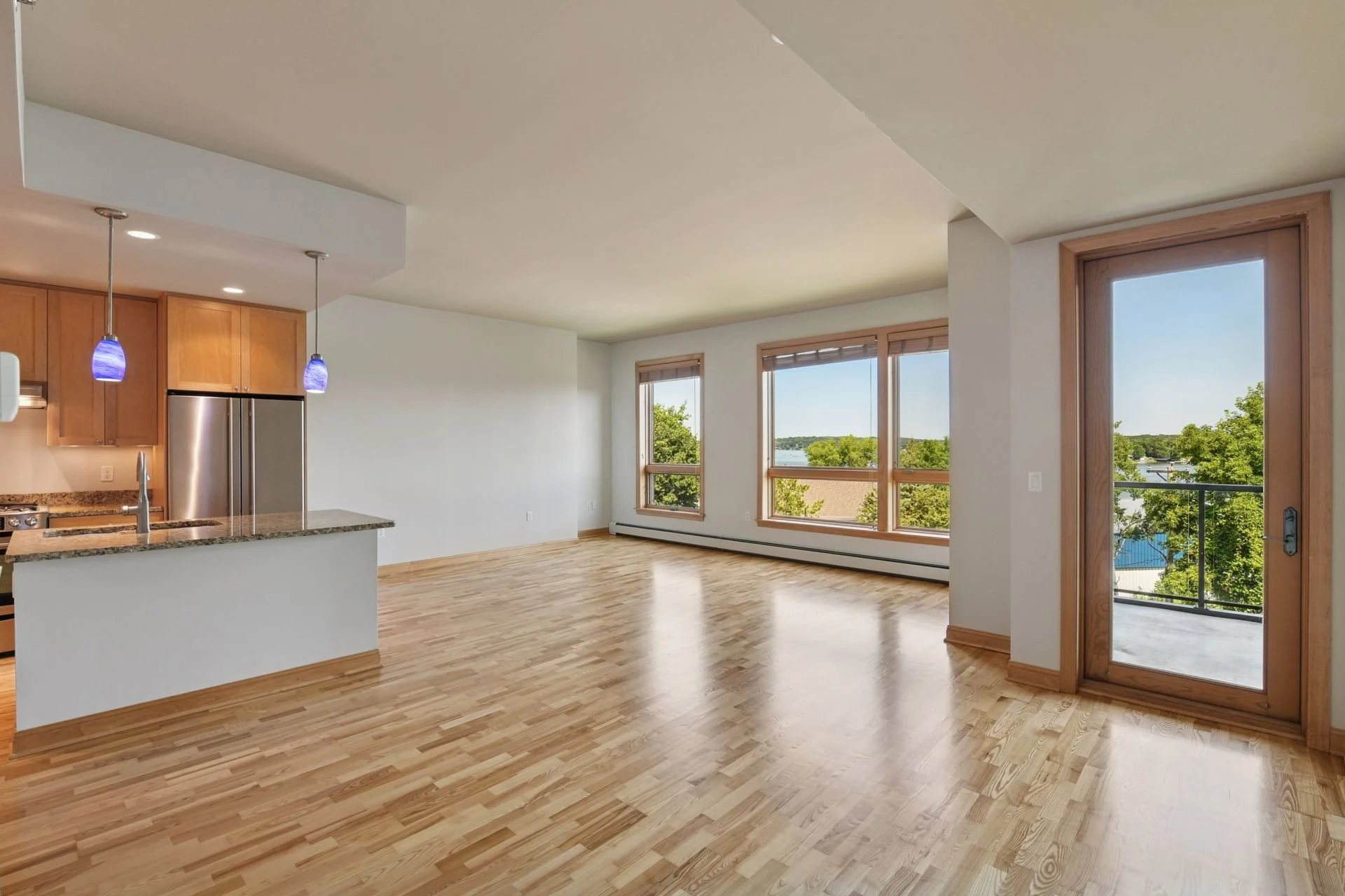 Living room with large windows, hardwood flooring, and adjacent kitchen with granite countertops and pendant lights.
