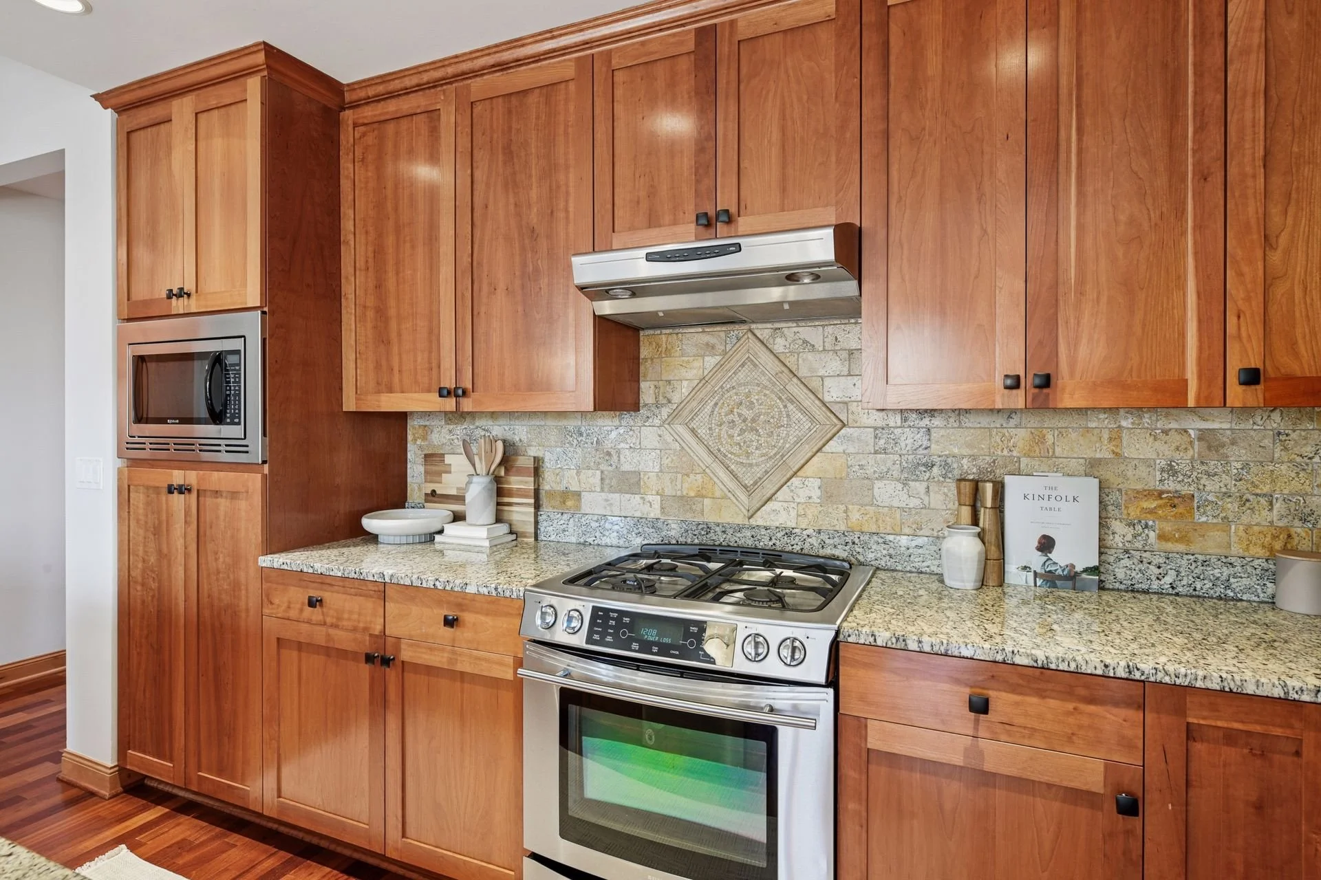 Wooden kitchen cabinets, granite countertops, a stainless steel oven with a gas stovetop, a wall-mounted microwave, and decorative wall tile.