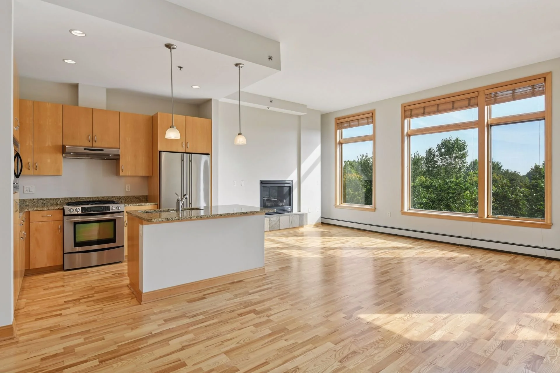 Empty open-concept living space with hardwood floors, large windows, a modern kitchen with granite countertops, wooden cabinets, stainless steel appliances, and a fireplace.