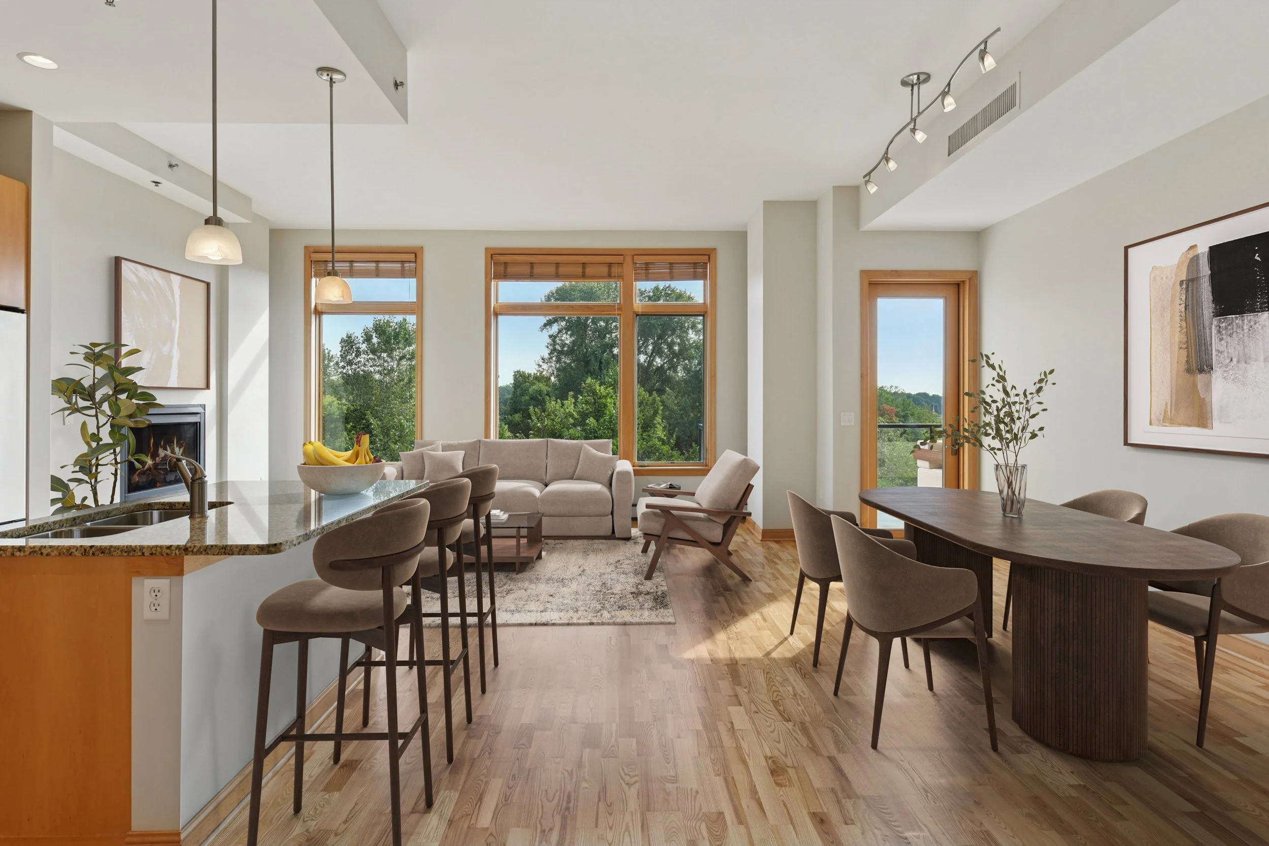 Bright living room with large windows showing trees outside, beige sofas, a modern dining table with chairs, hardwood floors, artwork on the walls, and a door leading outside.