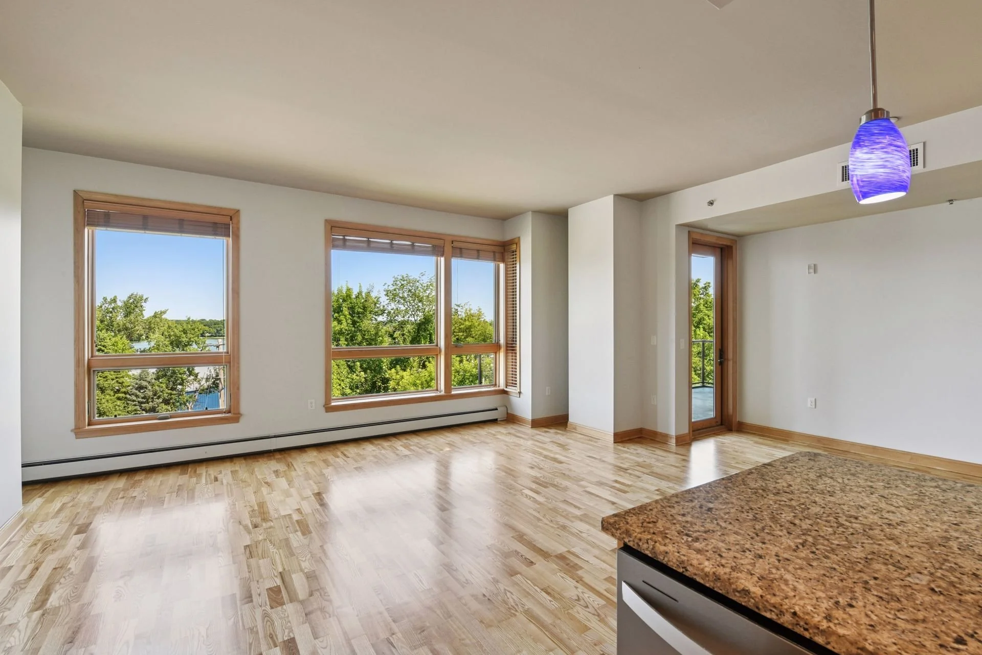 Living room with large windows revealing a view of green trees and a blue sky, hardwood floors, white walls, a door leading to a balcony, and a hanging blue pendant light.