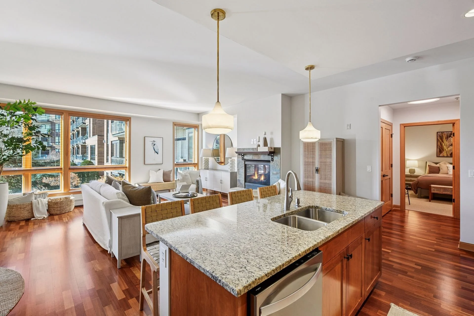 Open-concept living room and kitchen with hardwood floors, large windows, white walls, a fireplace, and a bedroom visible through an open doorway.