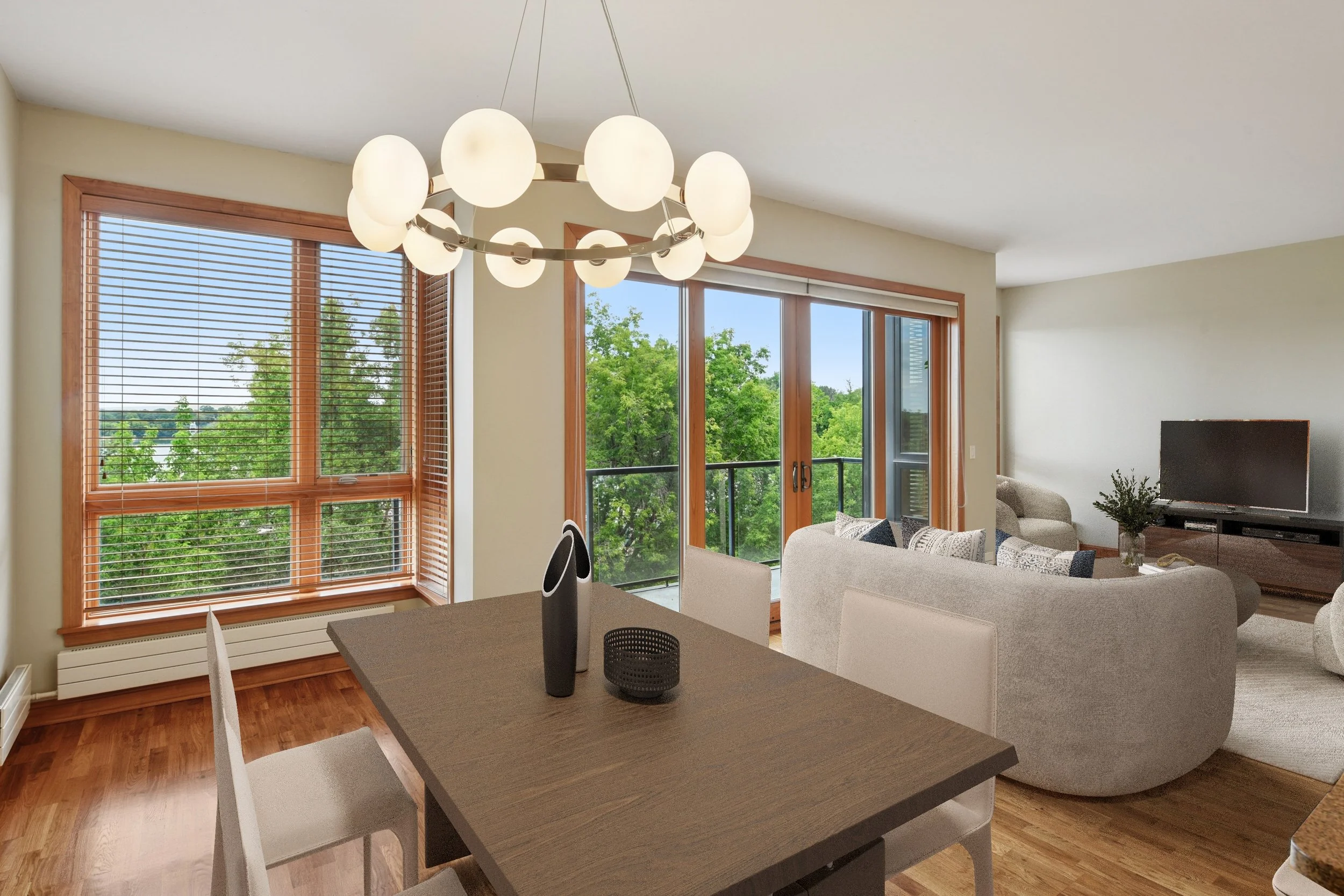 Living room with beige couches, wooden floor, large windows with blinds, sliding glass door leading to balcony, TV on a stand, and a dining table with chairs and a decorative object.