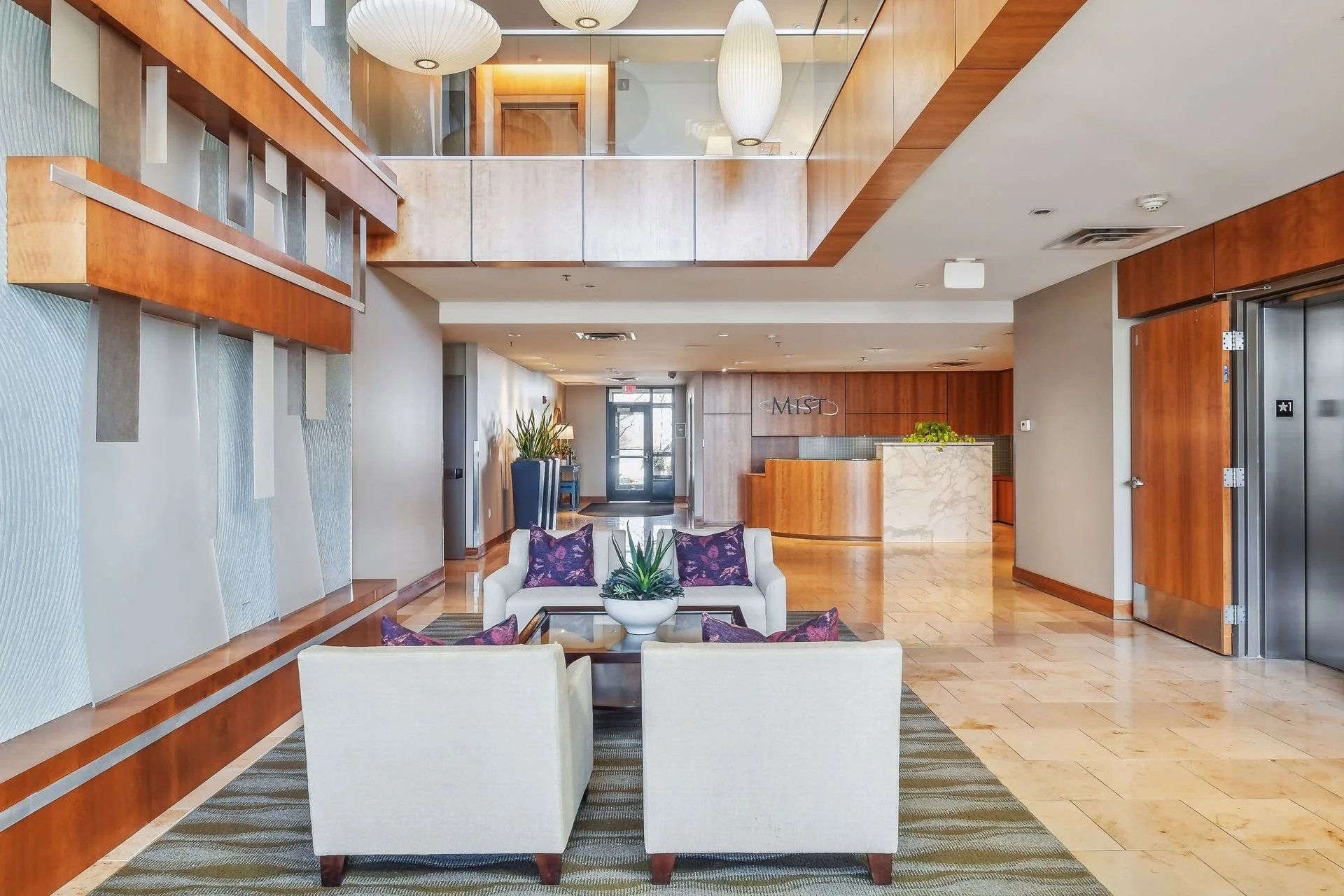 Modern lobby with white couches and purple pillows, a glass coffee table with a potted plant, wood-paneled walls, elevator doors on the right, and an reception desk in the background.