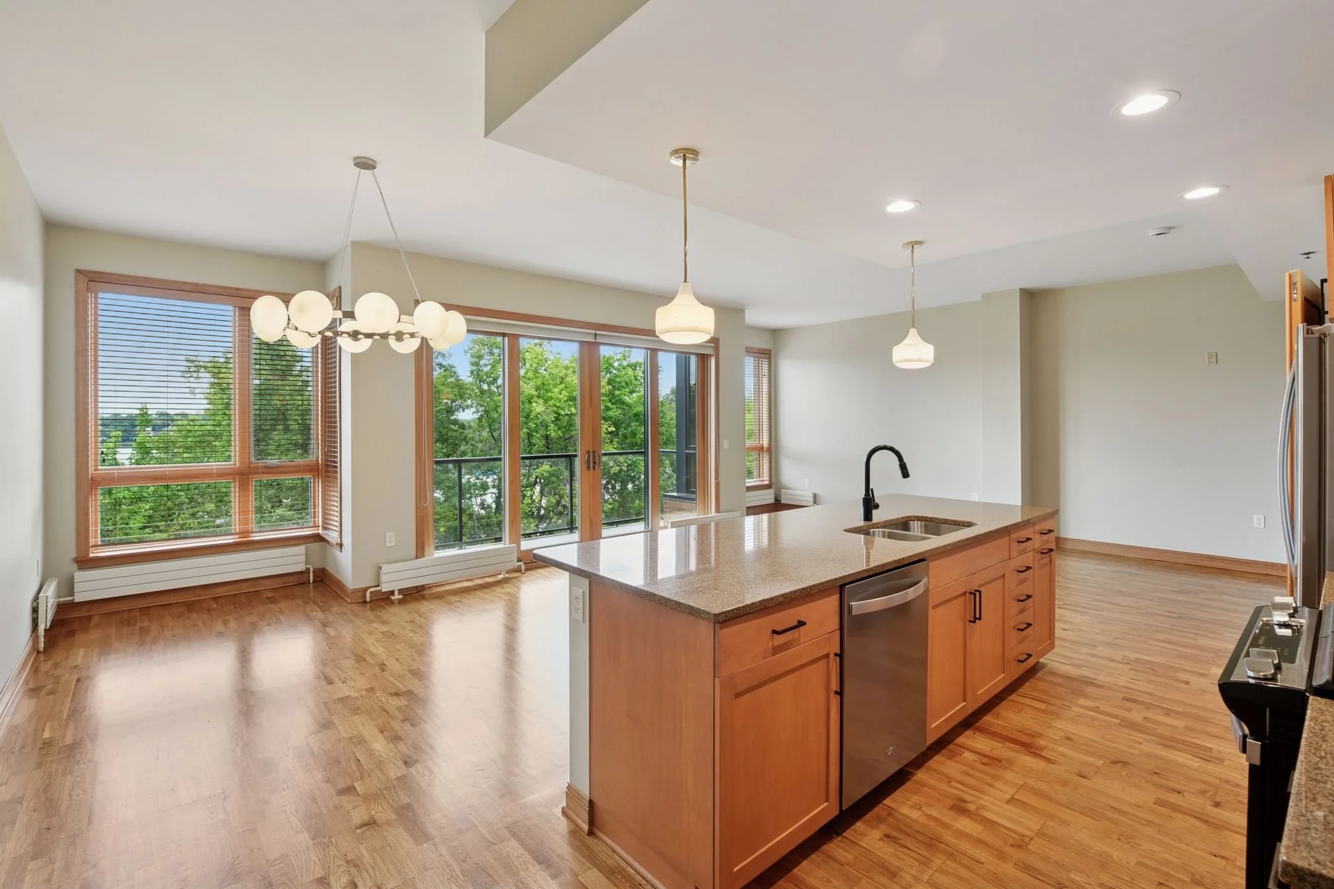 Open-plan kitchen and living area with hardwood floors, large windows, and a balcony with a view of green trees. The kitchen has a central island with a sink and modern appliances, pendant and chandelier lighting.