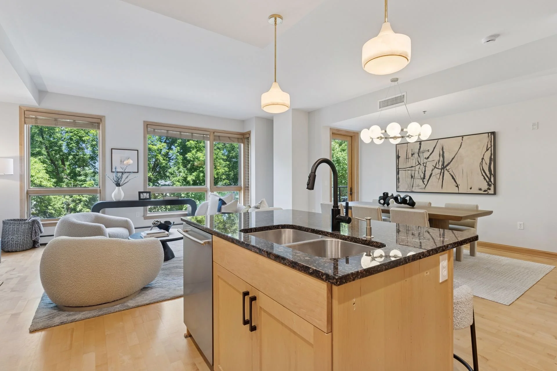 Open-concept living area with large windows, modern furniture, and a kitchen island with a dark granite countertop and sink. The space features modern light fixtures. There is outdoor greenery visible through the windows.