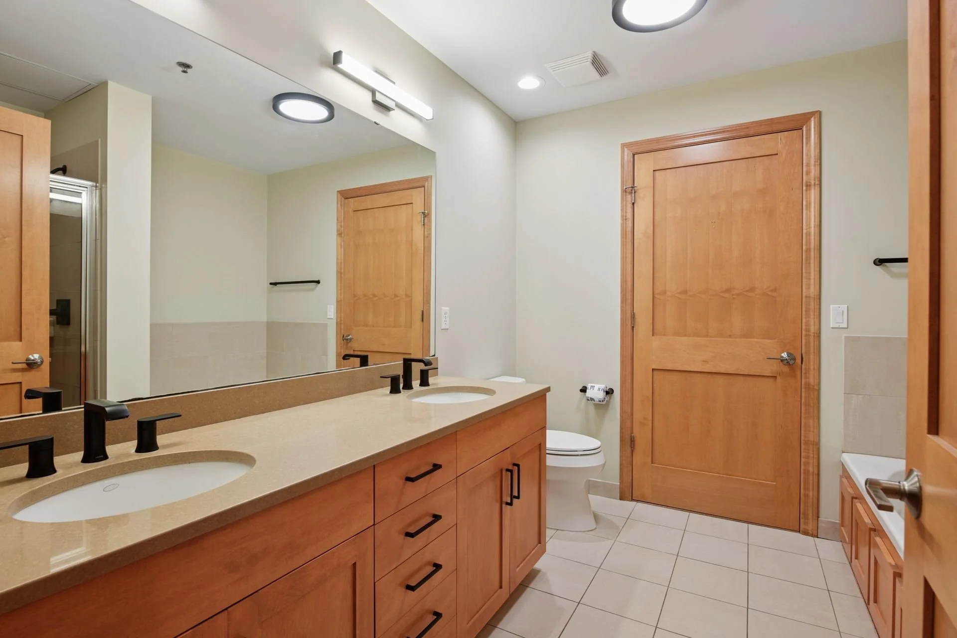 Bathroom with double vanity, wooden cabinets, black faucets, large mirror, toilet, and shower with glass door, neutral tile flooring and walls, and wooden door.