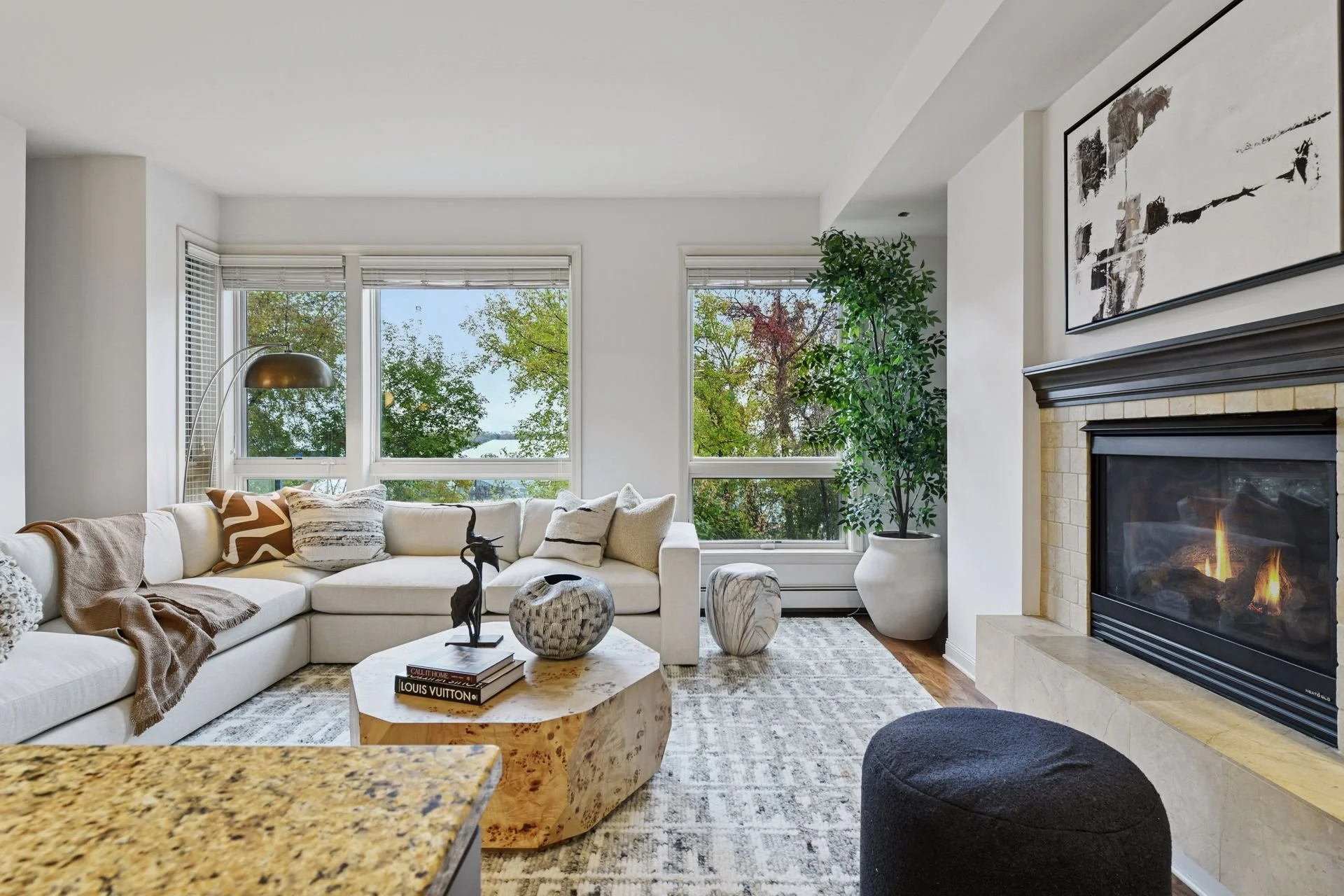 A bright living room featuring a fireplace and large windows showing trees outside.