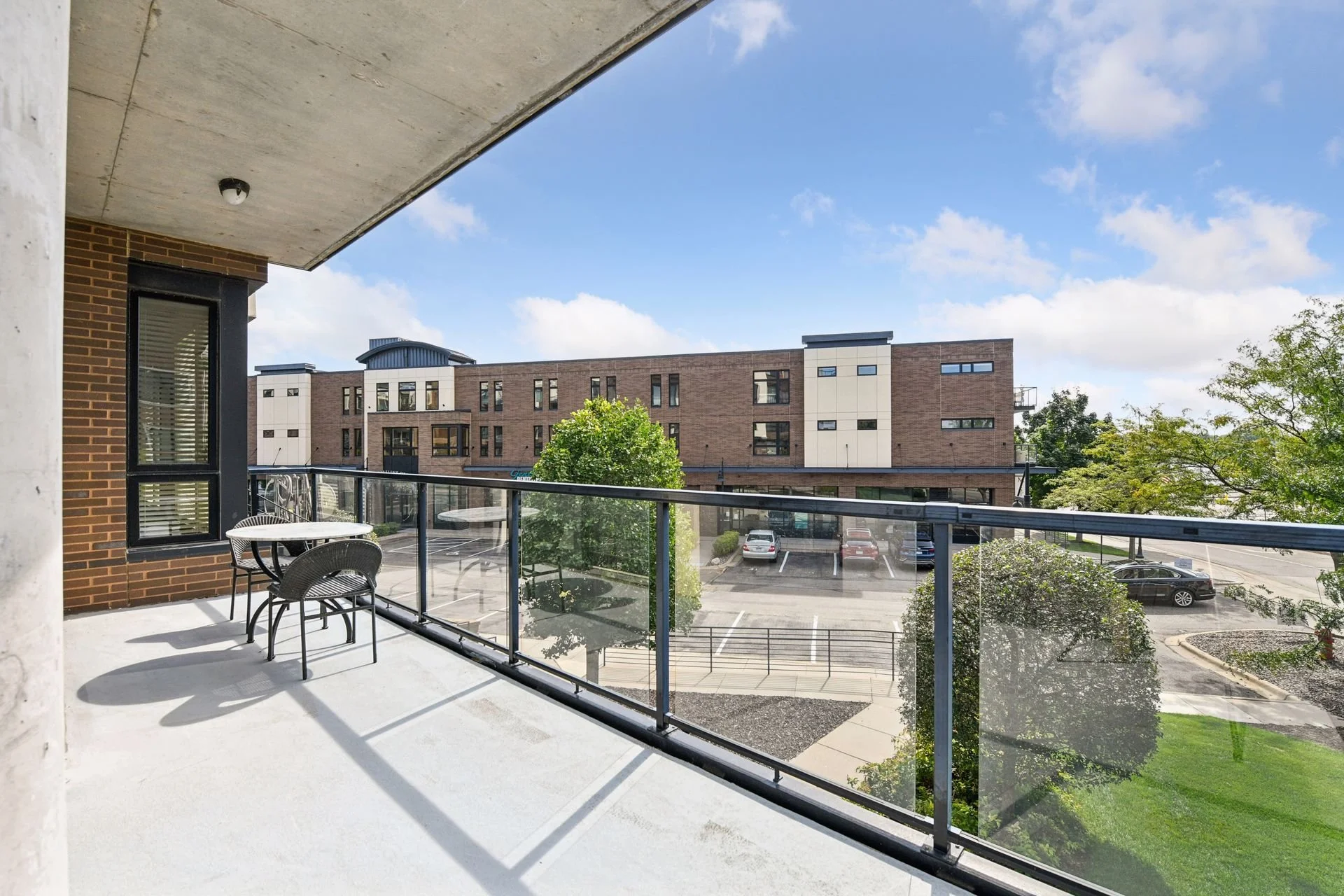 Balcony overlooking a parking lot and modern building with a brick facade, trees, and a blue sky with clouds.