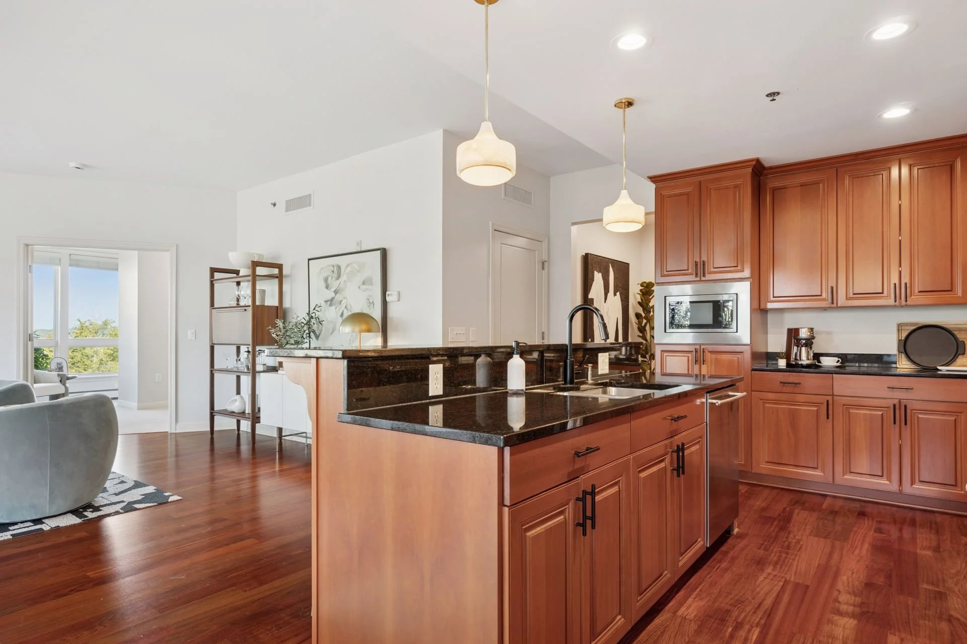 Open-concept kitchen with wooden cabinets, black granite countertops, and a spacious island. Stainless steel appliances, pendant lighting, and hardwood floors. A view into the living area with a large window.