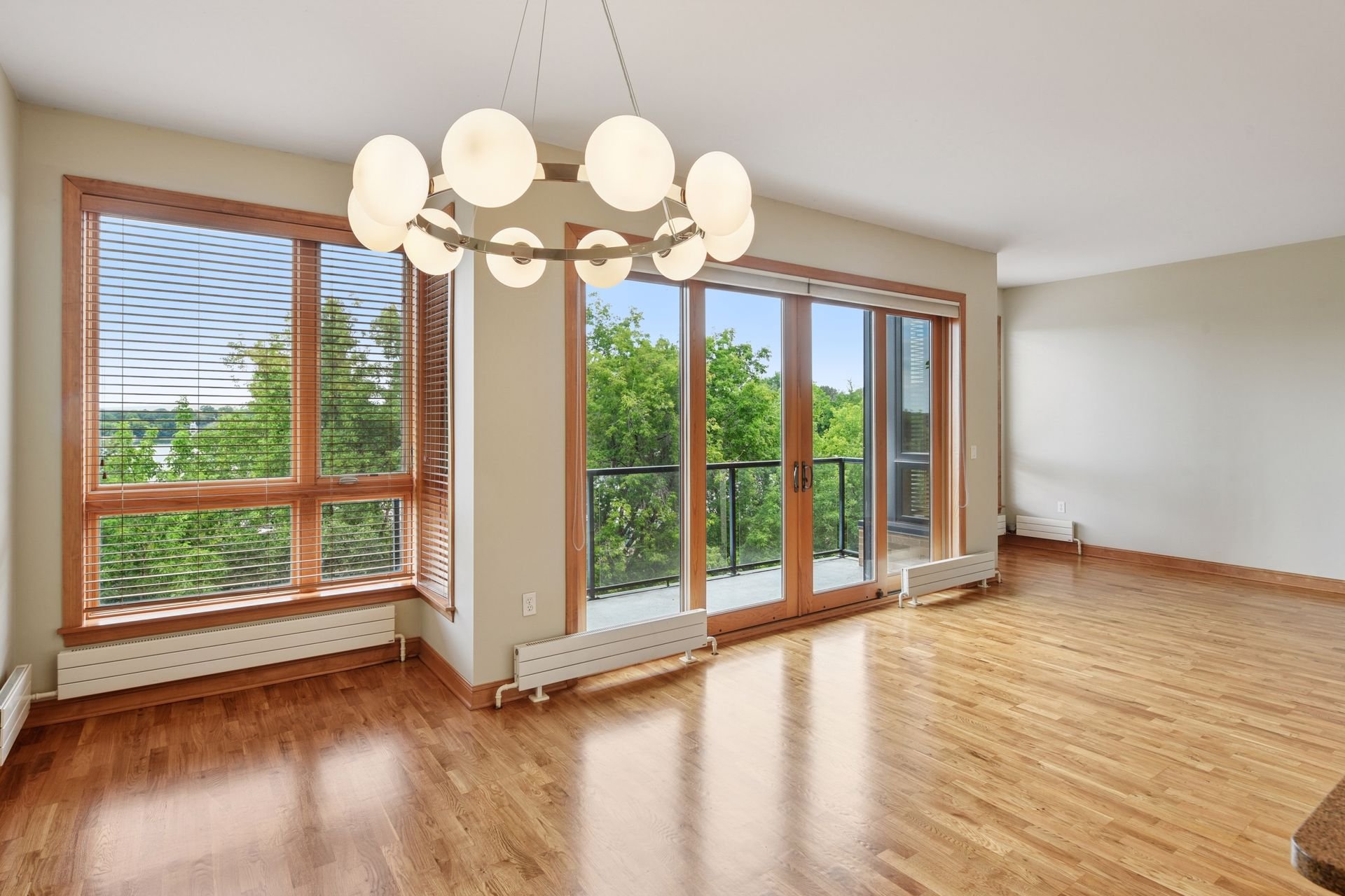 Empty living room with hardwood floors, large windows, a sliding glass door leading to a balcony, and modern lighting fixture.