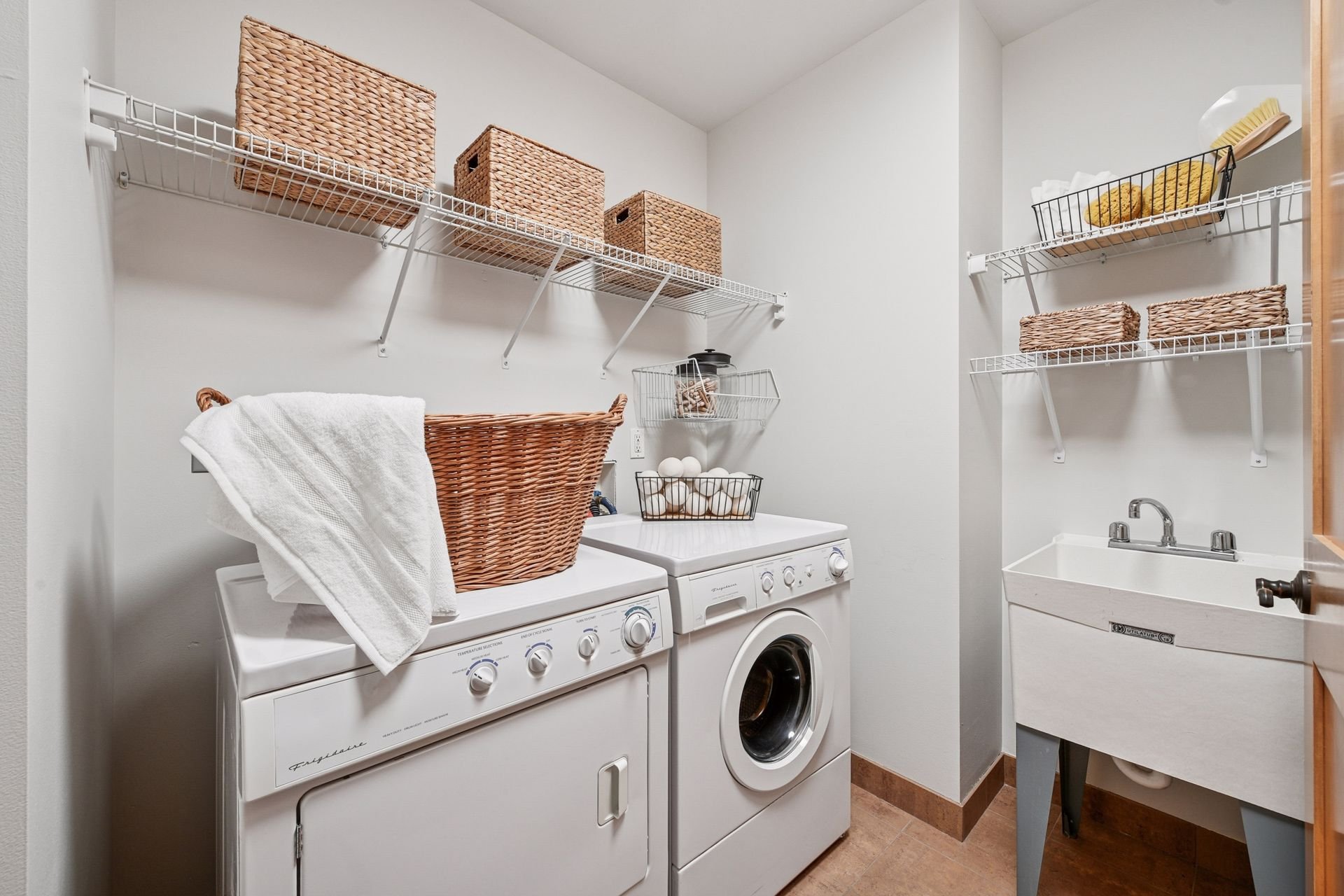 Laundry room with white washer and dryer, stored baskets on wire shelves, a laundry sink, and drying rack.