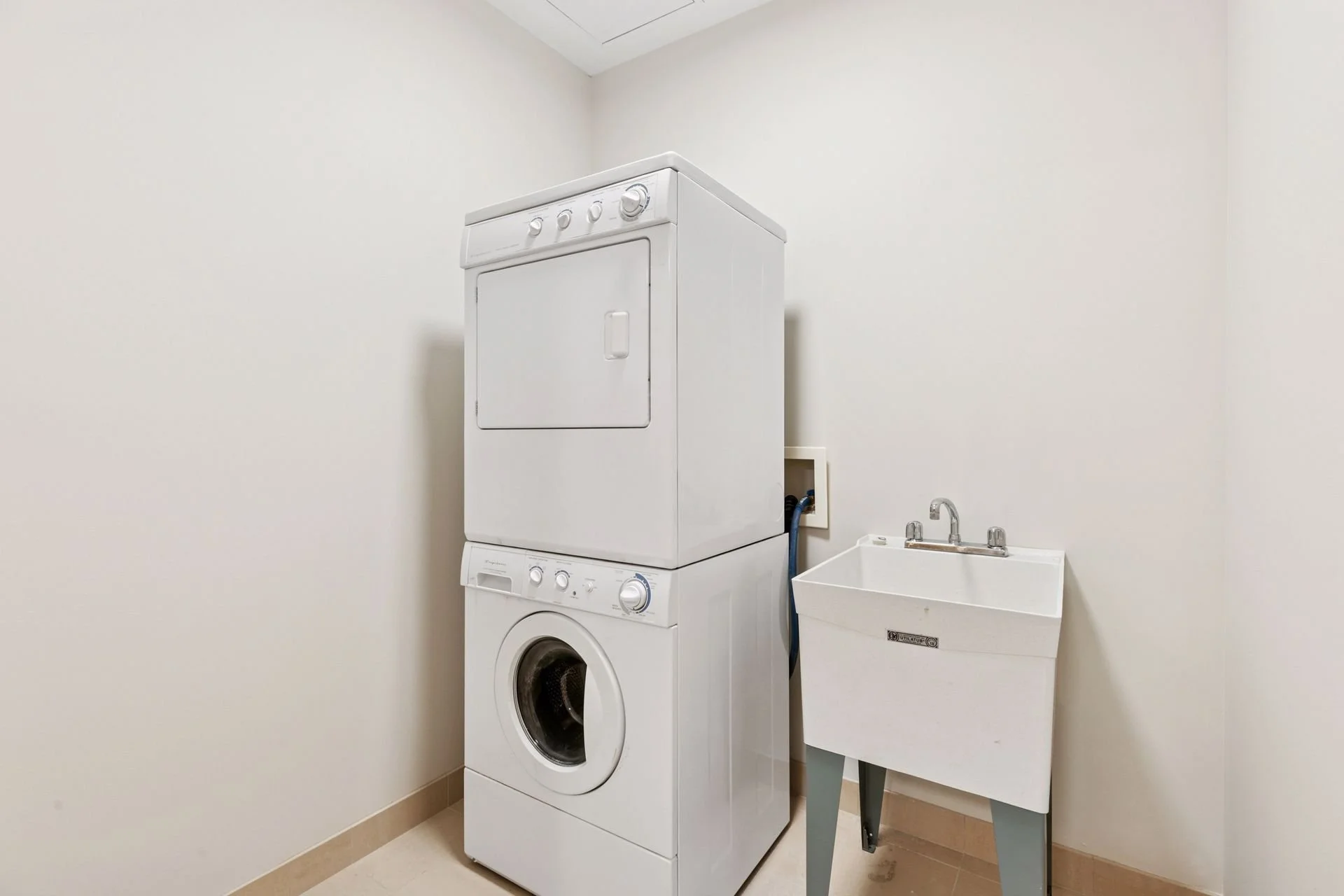 A white stacked washer and dryer unit in a laundry room corner, with a utility sink on gray legs nearby.