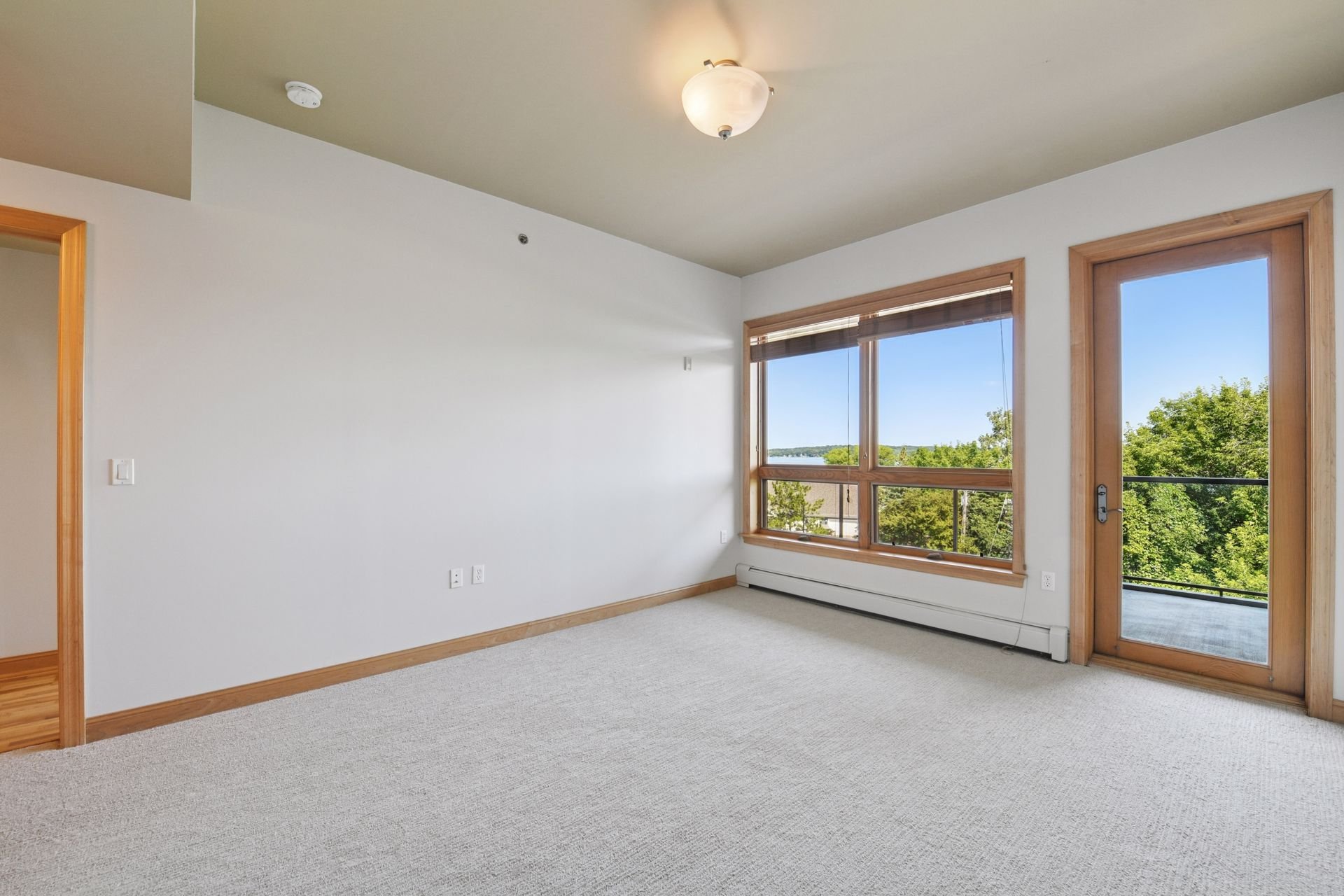 Large bedroom with large window and glass door leading to balcony, white walls, beige carpet, and wooden trim.