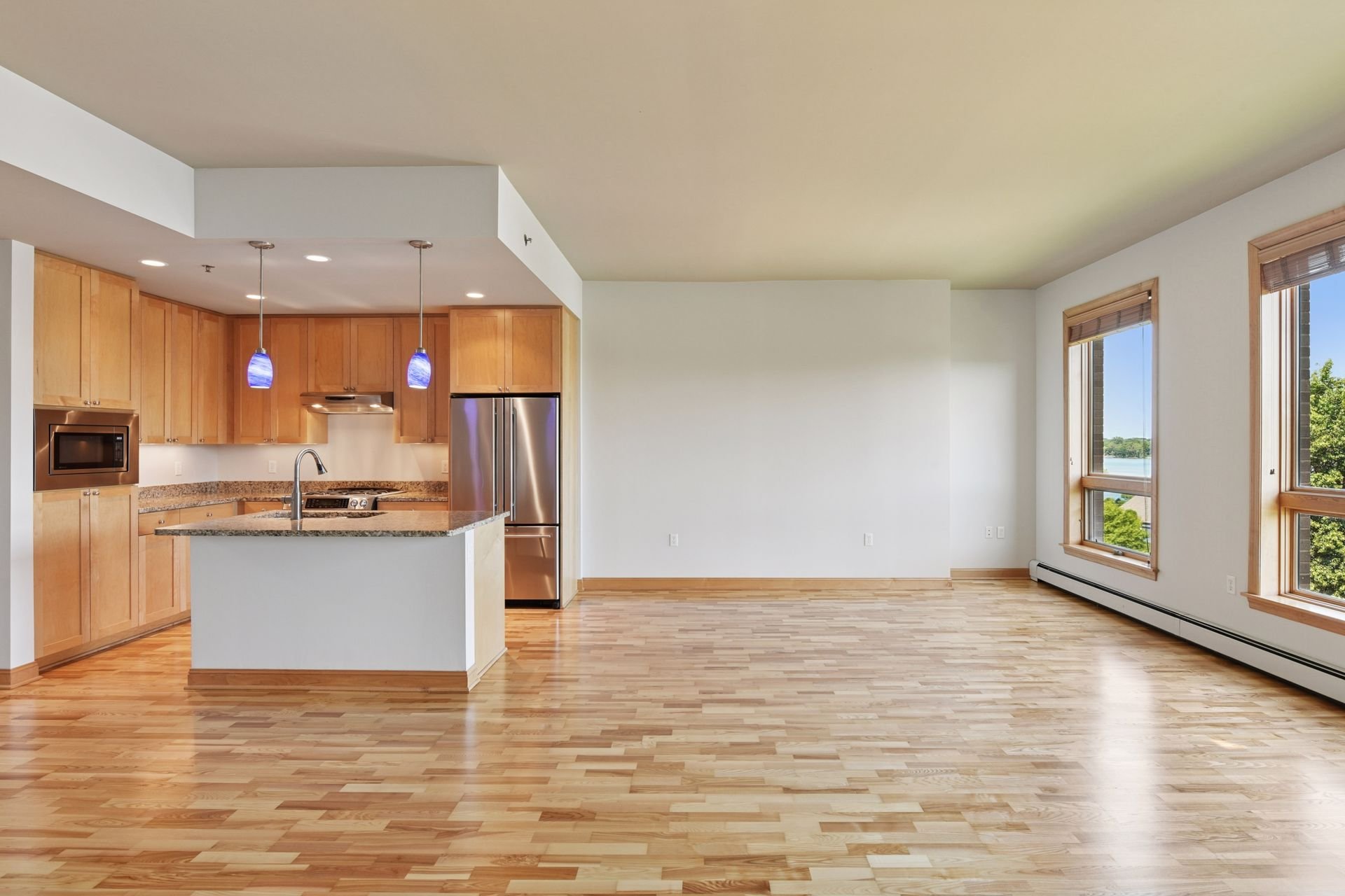 Condo with open floor plan, hardwood floors, large windows, and a kitchen with wooden cabinets, granite countertops, stainless steel appliances, and pendant lighting.
