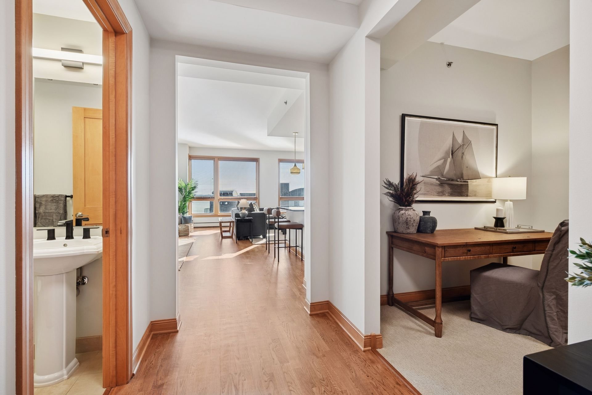 Hallway leading to an open living space with large windows, a sitting area, and a kitchen. To the right, a small office nook. To the left, a bathroom.