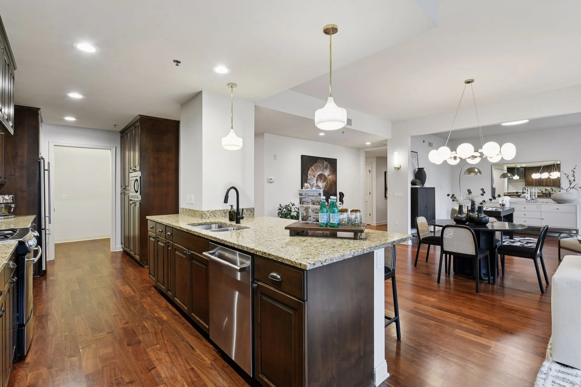 Open floor plan of a modern apartment featuring a kitchen with granite countertops and dark wood cabinets, a dining area, hardwood floors, and contemporary lighting fixtures.