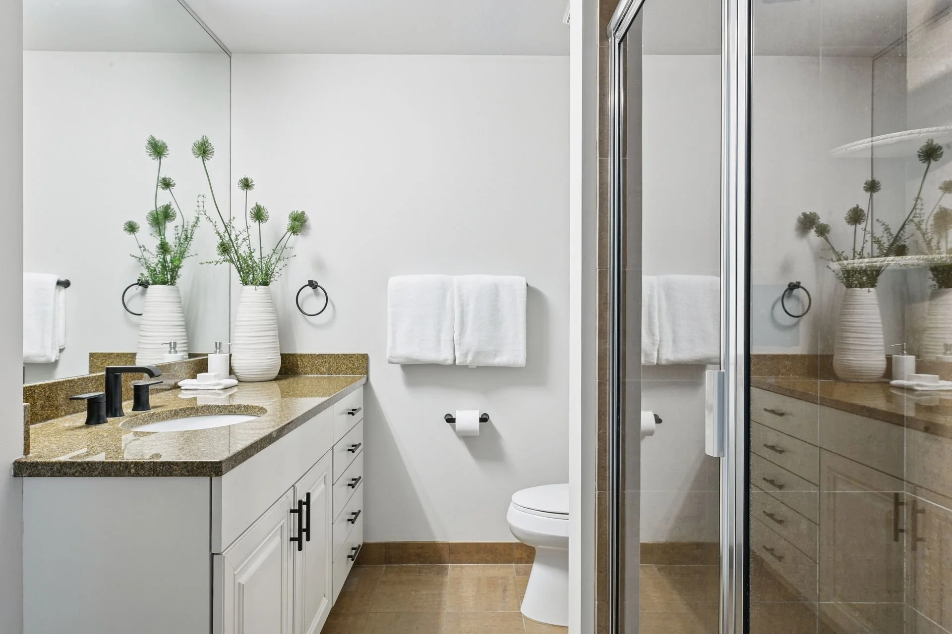Bathroom with white walls, a vanity with a gold countertop, a mirror, a towel ring, a toilet, and a glass shower enclosure with brown tiles.