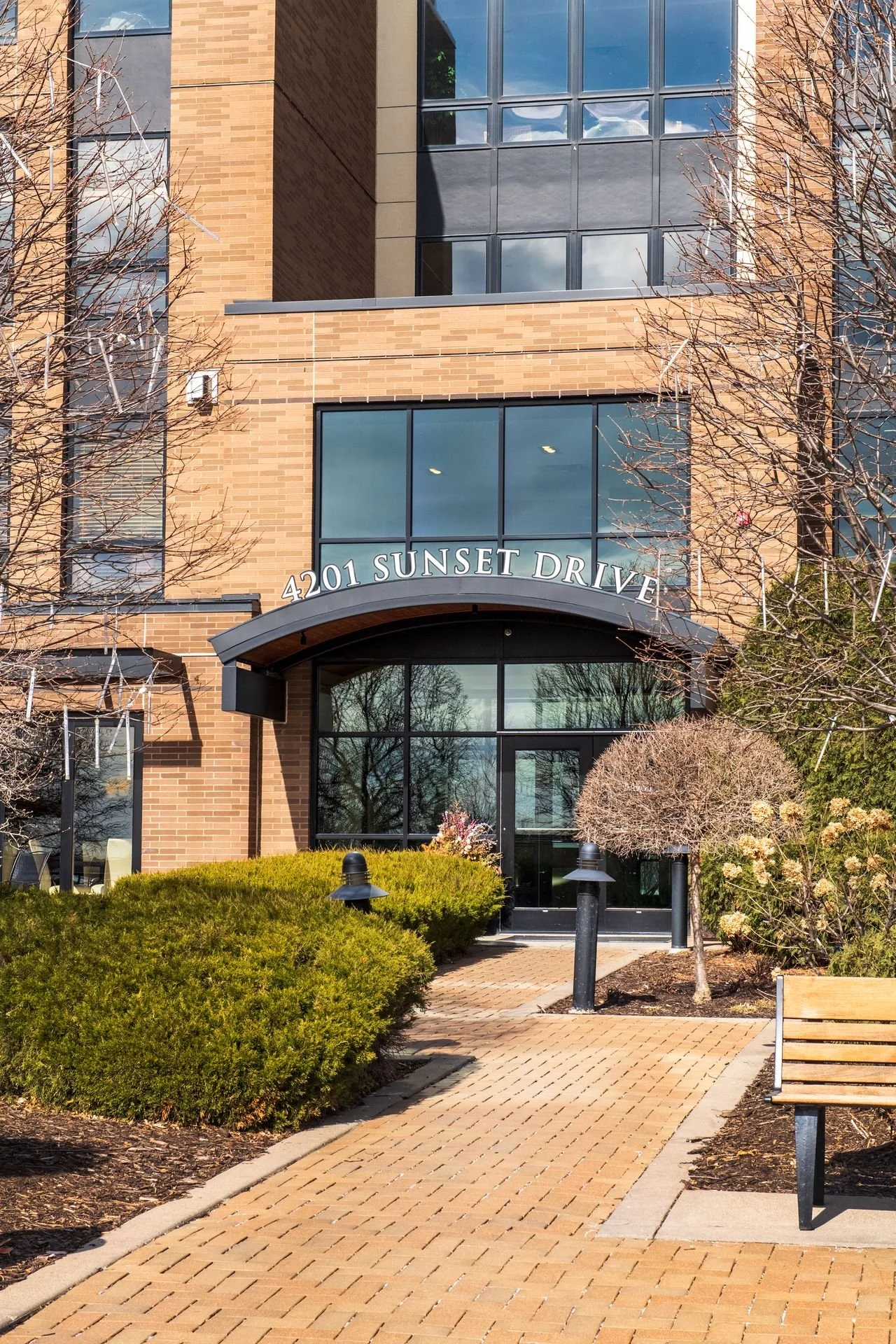 Front entrance of a modern building with brick and glass facade, displaying the address 4201 Sunset Drive. The pathway leading to the entrance is paved with bricks, flanked by trimmed bushes and small trees, with benches and outdoor lighting.