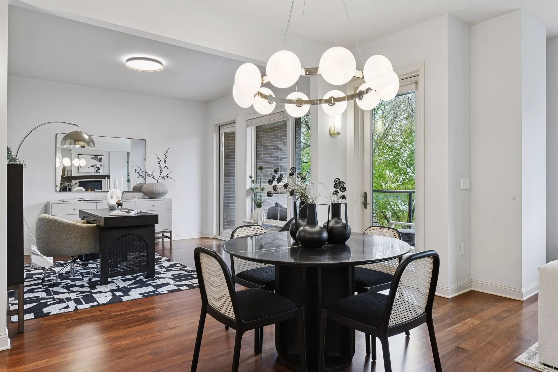 Modern dining and living room with hardwood floors, and large windows with a view of green trees.