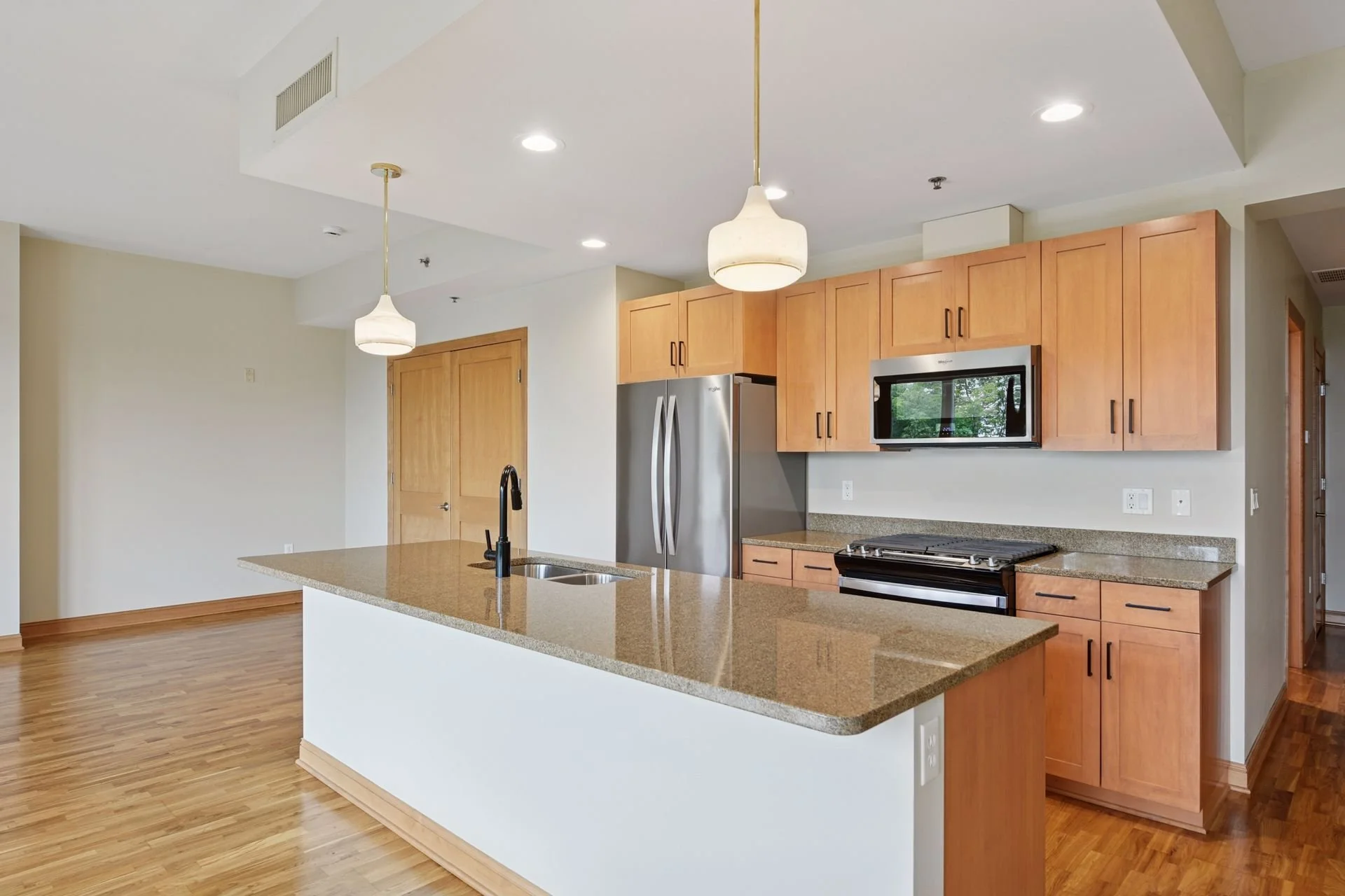 Modern kitchen with wooden cabinets, stainless steel refrigerator and microwave, granite countertop island with sink, and hardwood floors.