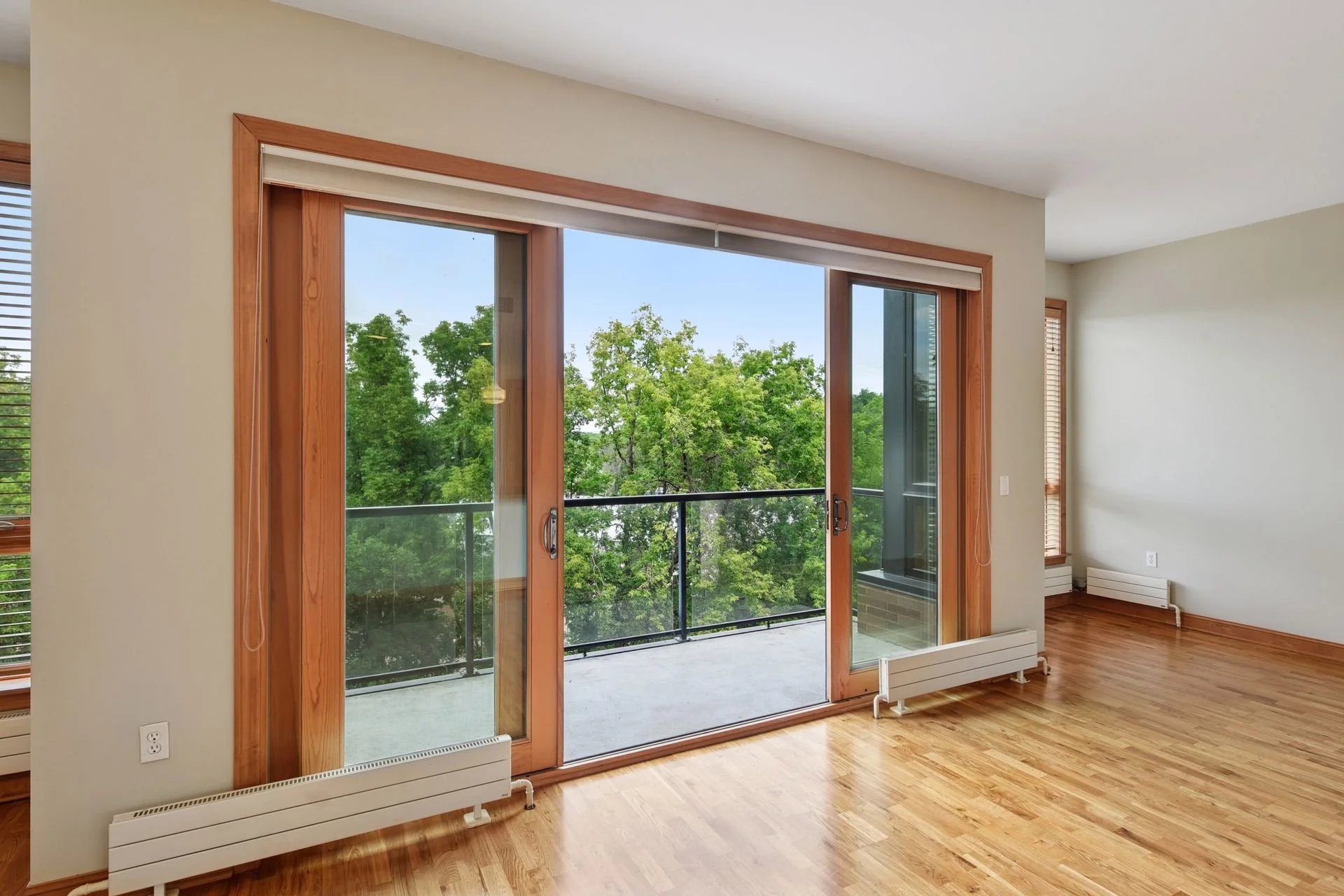 Empty room with hardwood floors, large glass sliding door leading to a balcony with greenery outside, and windows with wooden frames.
