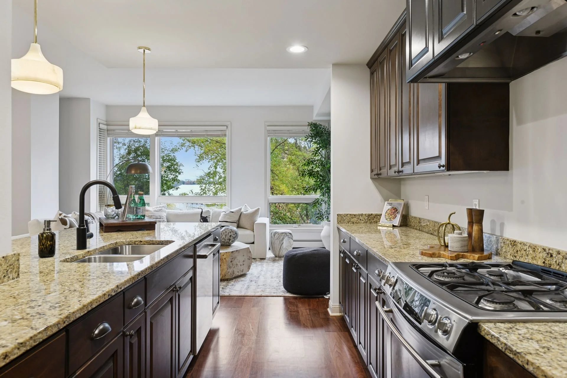 Kitchen with granite countertops, dark wood cabinets, stainless steel appliances, and a view of a sunny living room with large windows.