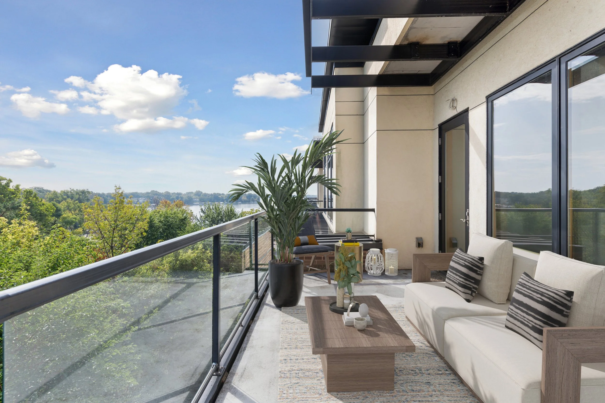 Balcony with glass railing, outdoor furniture, potted plant, scenic view of trees and river, blue sky with clouds.
