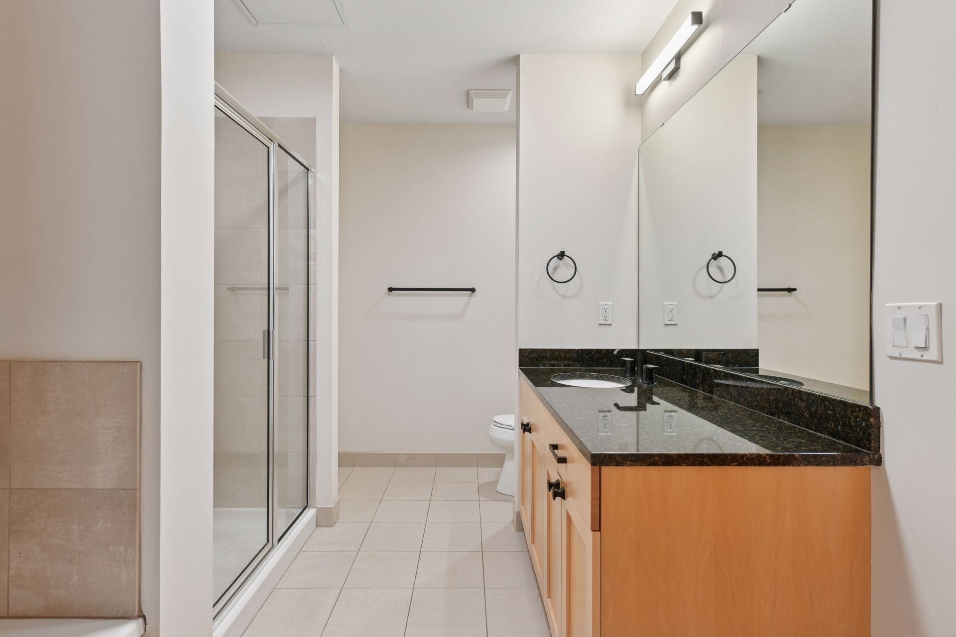 Bathroom with a glass shower, a toilet, a large mirror, black countertop with a sink, and beige tiles.