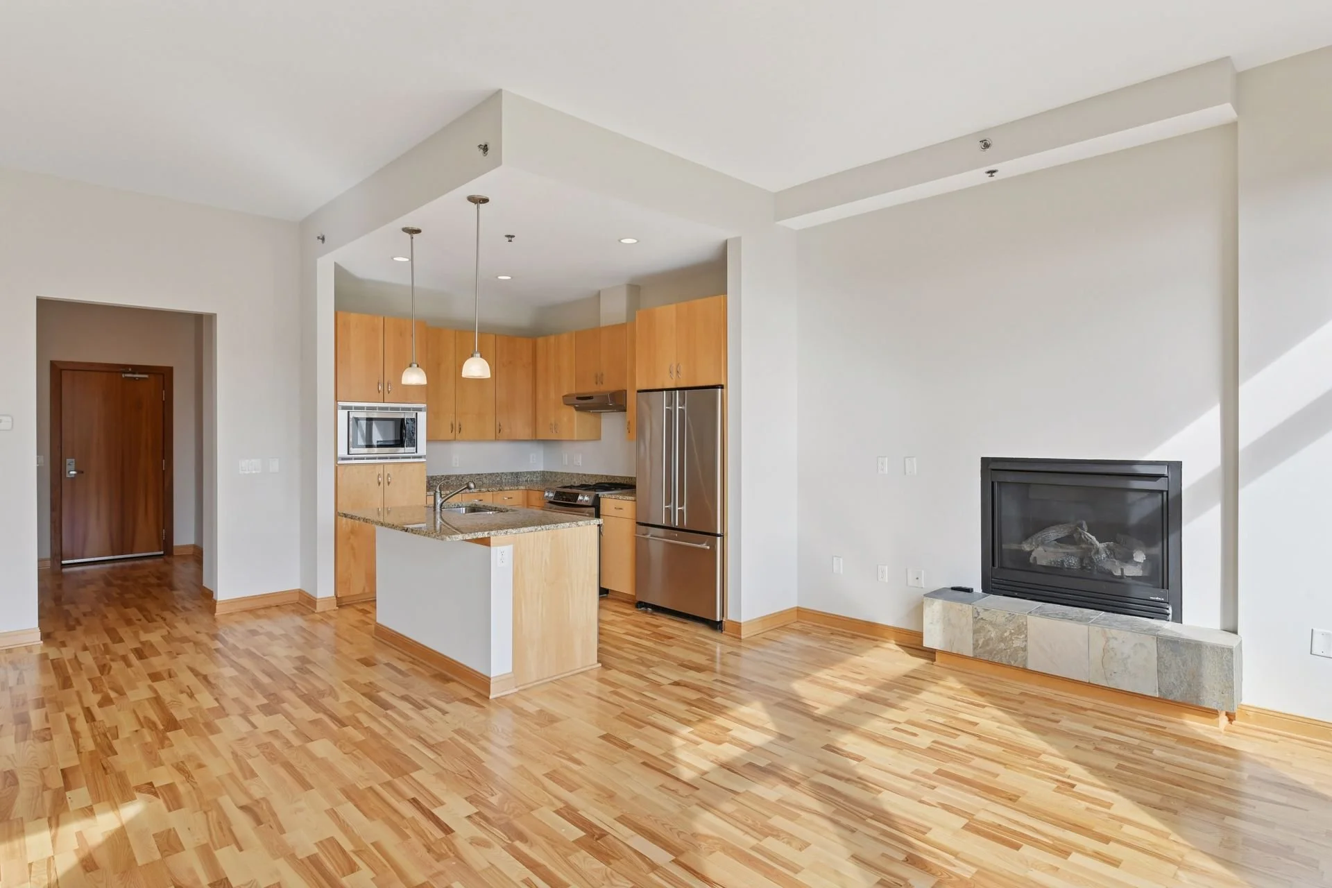 Empty living room and kitchen area with hardwood floors, white walls, a fireplace, and wooden cabinetry.