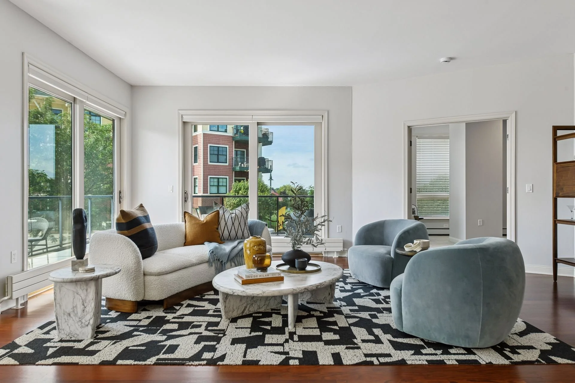 Living room with large windows showing apartment building and balcony outside.