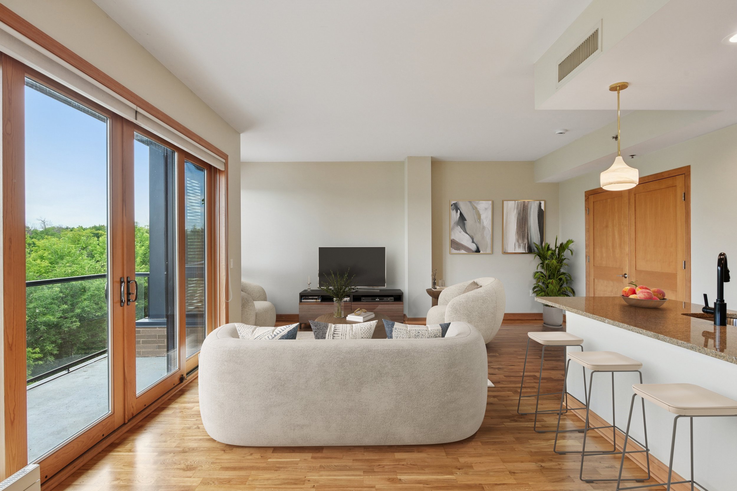 Bright living room with large sliding glass doors, a white curved sofa, a TV on a stand, two armchairs, a coffee table, wall art, a plant, and a kitchen counter with three barstools.