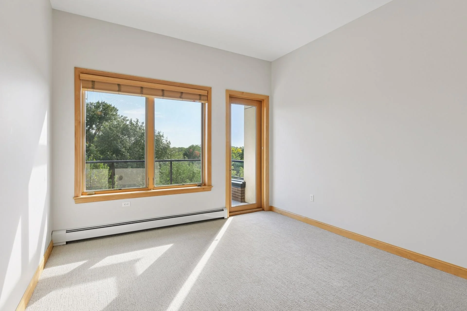 Empty room with large window and balcony door, white walls, wooden trim, beige carpet, and green trees outside.