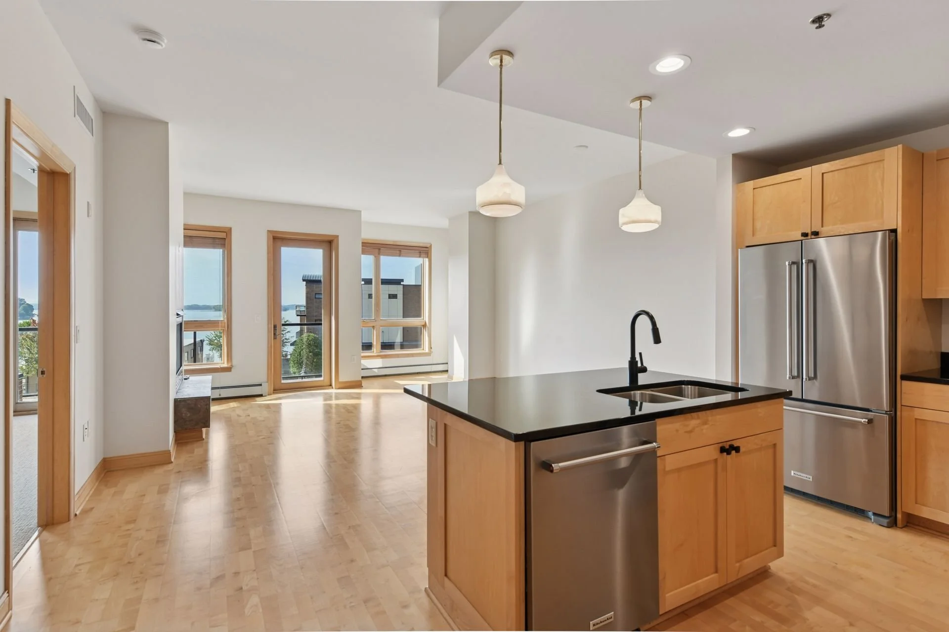 Modern kitchen with wooden cabinets, stainless steel refrigerator, black countertop island, black faucet, two pendant lights, and hardwood flooring. Large windows in the living area with view of buildings and trees.