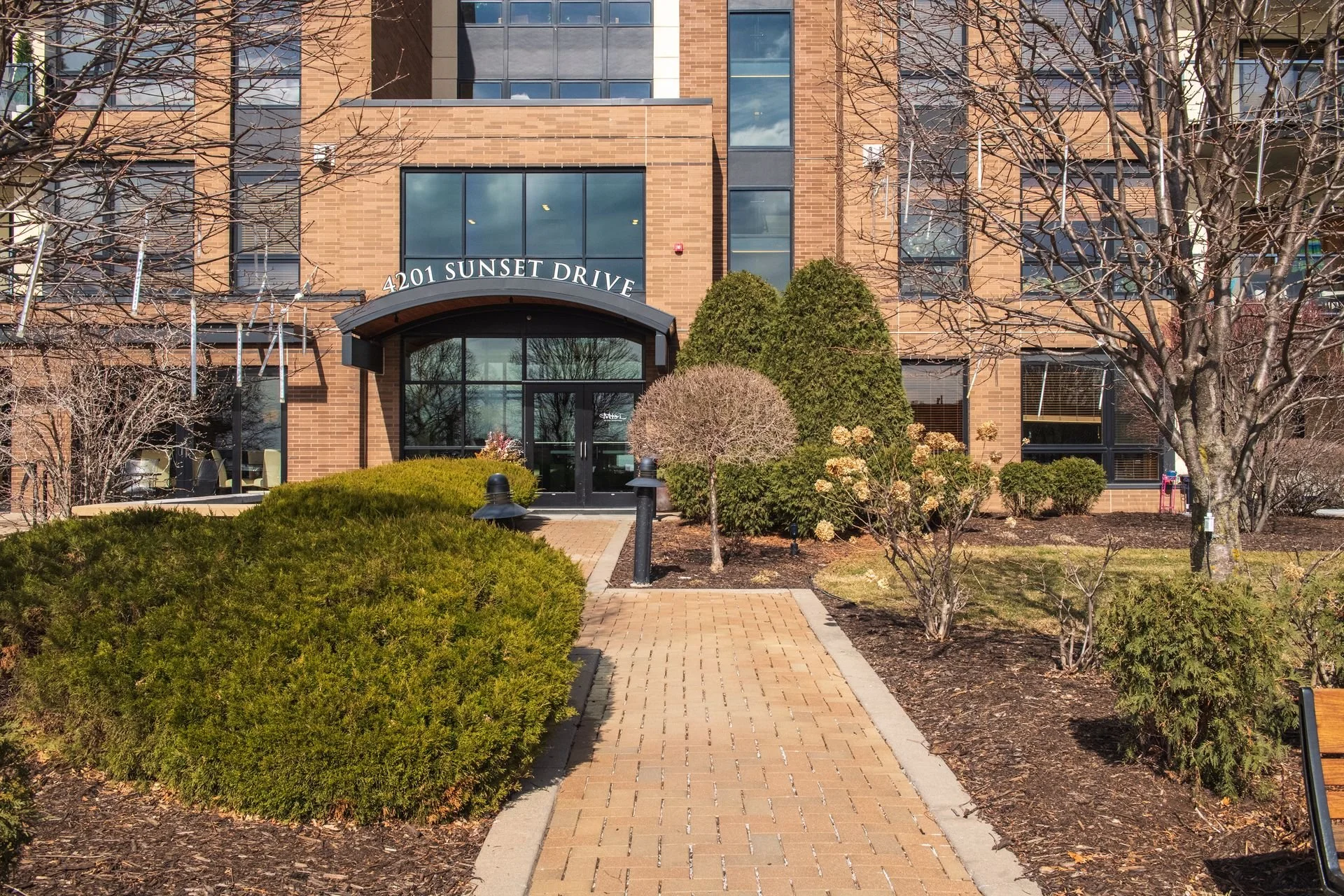 Entrance to a residential building at 4201 Sunset Drive, featuring a brick facade, black awning, a pathway, and landscaped bushes and trees.