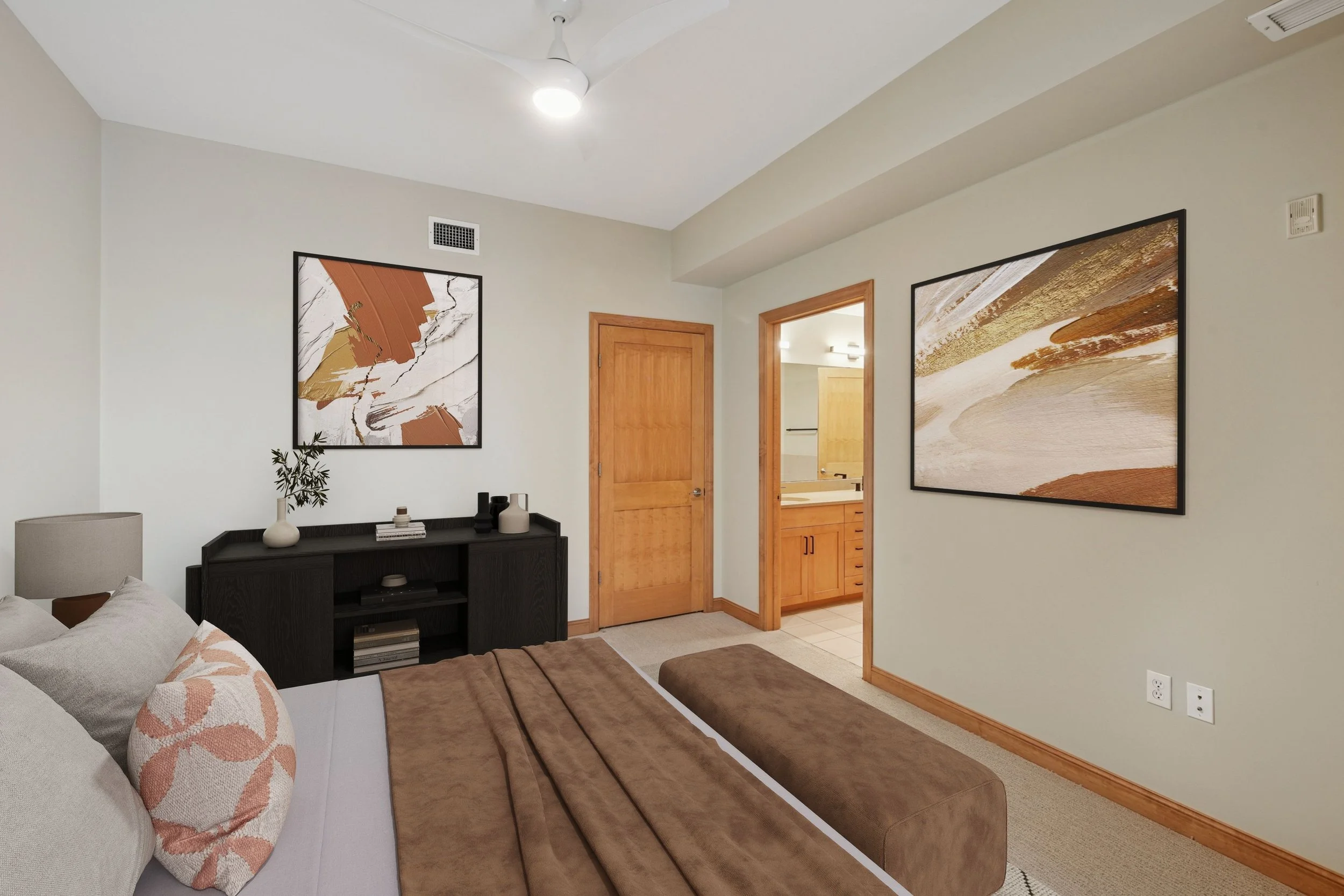 A bedroom with a bed, decorative pillows, a black dresser with vases and books, wall art, a doorway to a bathroom, and wall outlets.