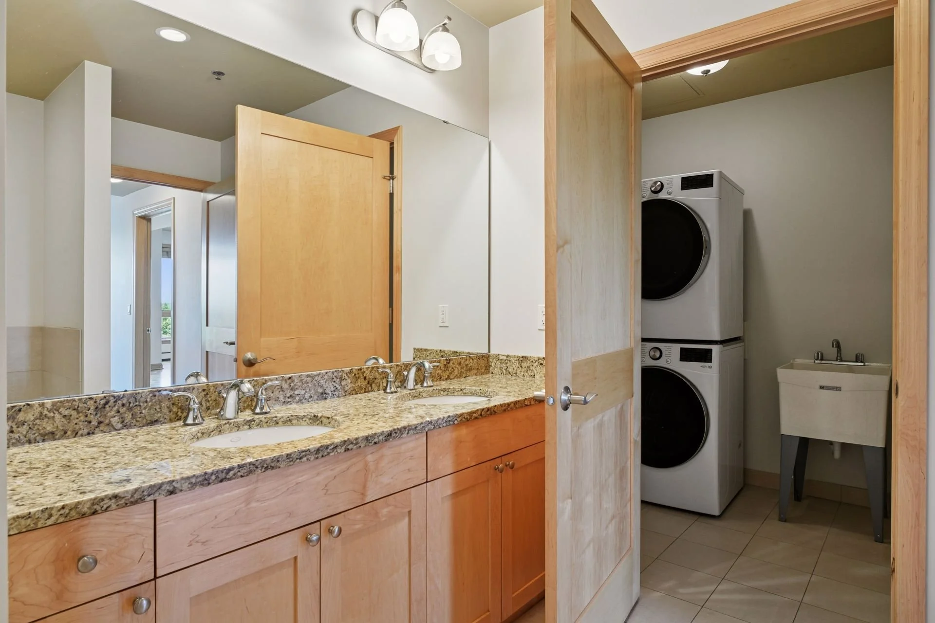 Bathroom with double sink granite countertop, wooden cabinets, mirror, washing machine, dryer, laundry sink, and door.