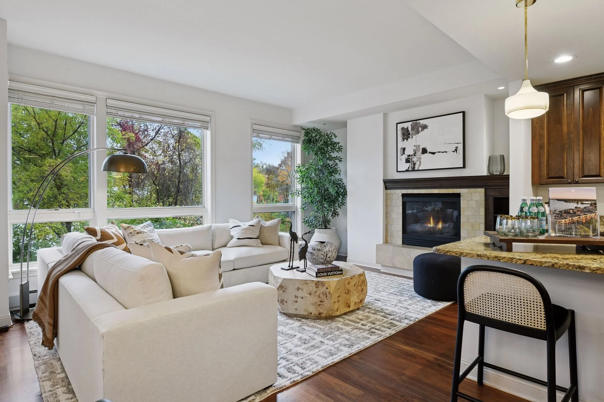 Modern living room with large windows with views of green trees, a fireplace, a spacious island, and hardwood floors.