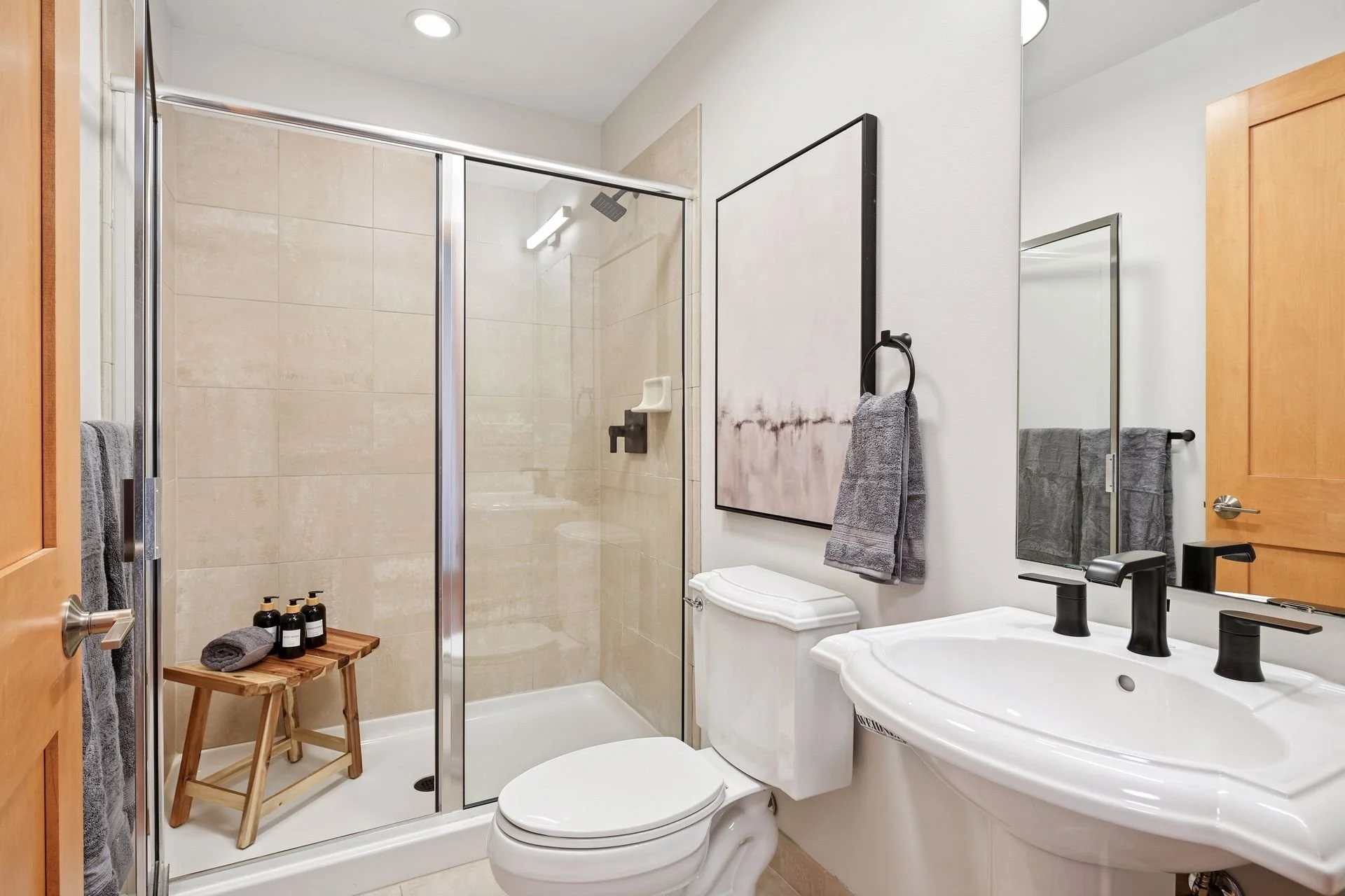 Modern bathroom with a walk-in shower, toilet, and sink. The shower has glass doors and beige tile walls. A large mirror is above the sink, and towels hang on the wall. The room has a neutral color scheme with natural wood accents.