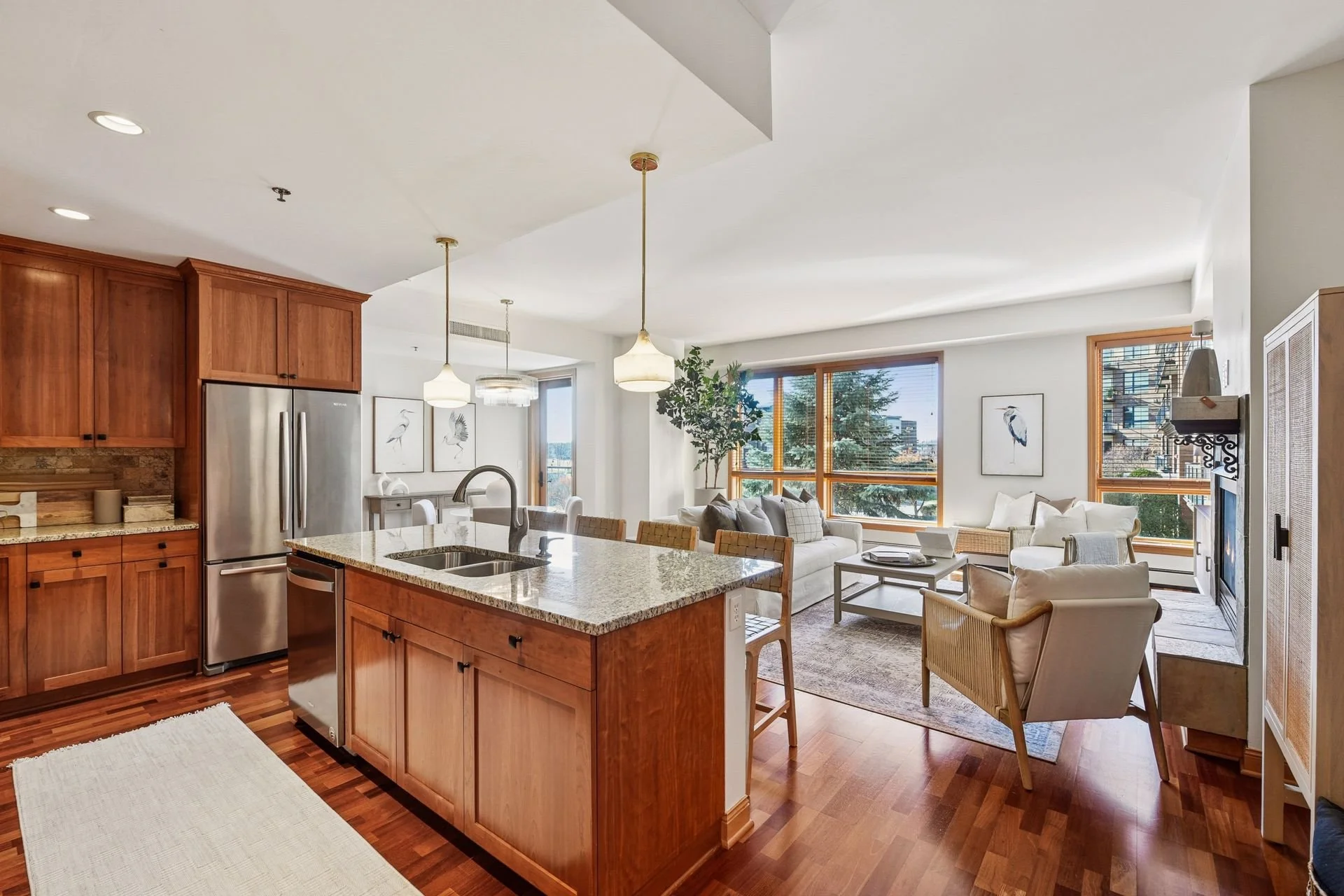 Open-concept living room and kitchen with wooden cabinets, granite countertops, stainless steel refrigerator, seating area with large windows, and a fireplace.