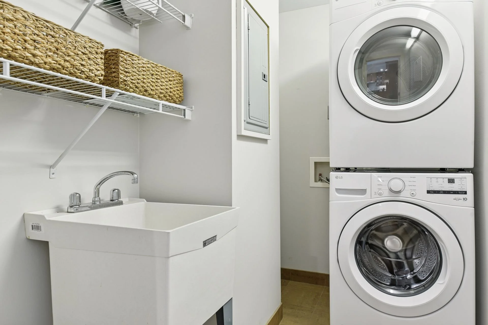 Laundry room with stacked washer and dryer, utility sink, wall-mounted wire shelf with baskets, and a wall-mounted electrical panel.