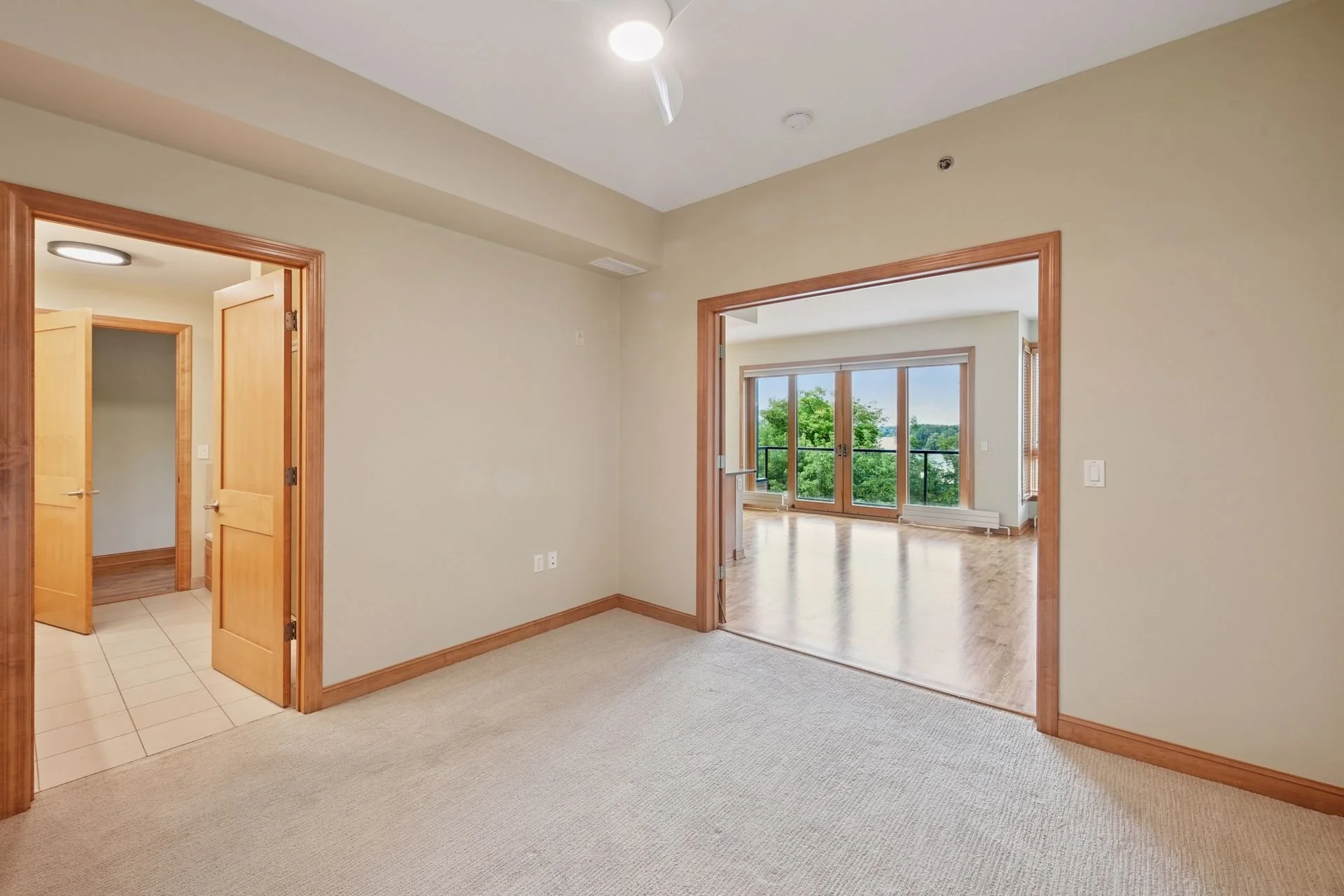 Empty room with beige carpet, beige walls, wooden doorframes, and view into living area with large windows and a balcony.