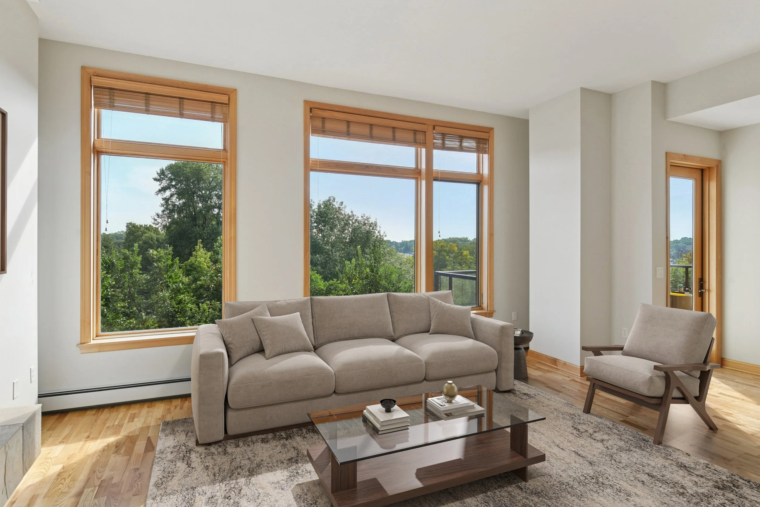 Living room with a beige sofa, a wooden armchair, a glass coffee table with books and decorative items, large windows with wooden frames and blinds, and a view of green trees outside.