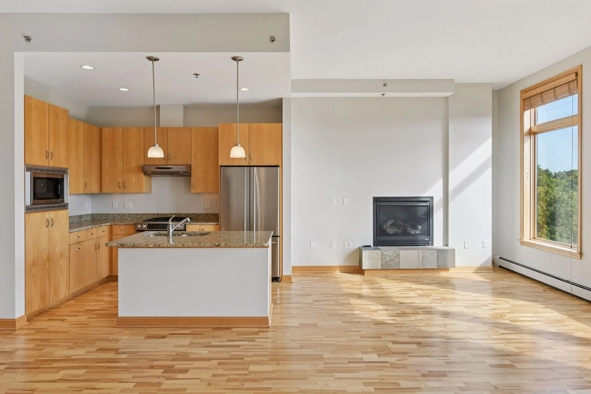 Open-concept living area with kitchen featuring wooden cabinets, granite countertops, and a central island; living room has a fireplace with a stone base, large windows with wooden trim, and hardwood flooring.